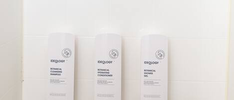 Un baño con ducha, tres botellas de champú, acondicionador y gel de ducha Ideology botanical, y un grifo moderno.