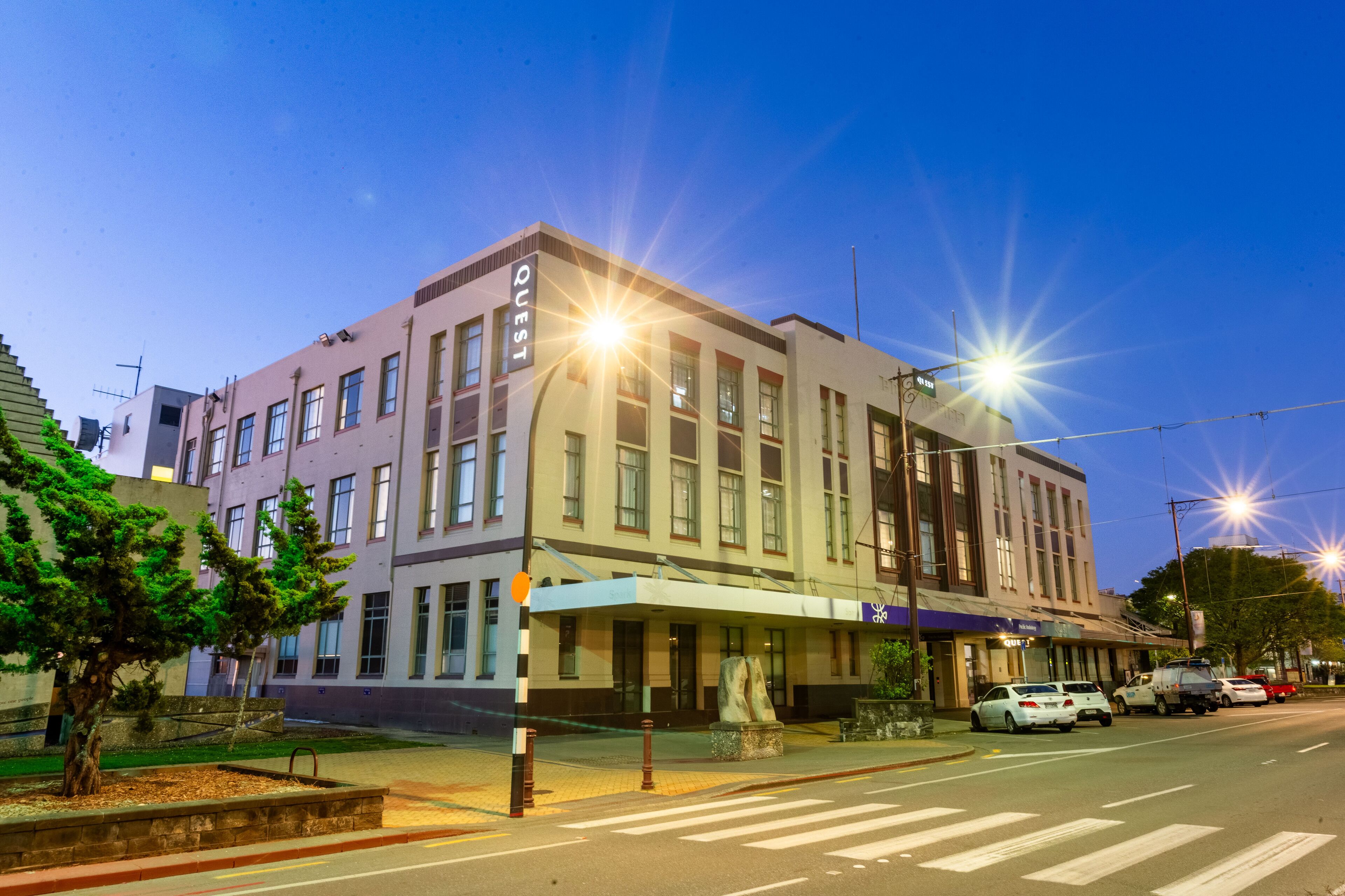 A multi-story building with a 'QUEST' sign, a covered entrance, and a parking lot with cars.
