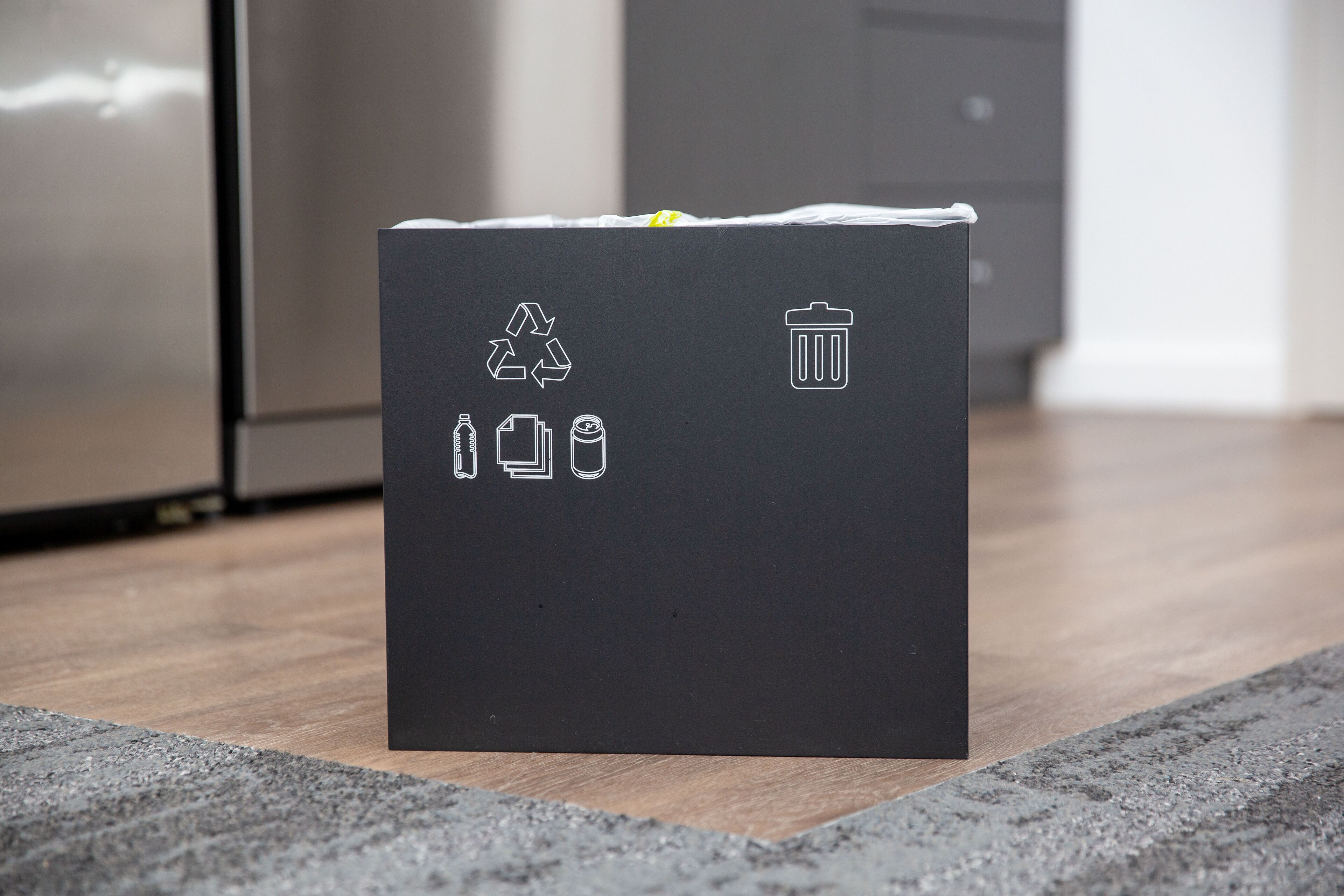 Une poubelle de recyclage noire, ornée de symboles de recyclage et de déchets, est posée sur un plancher en bois.