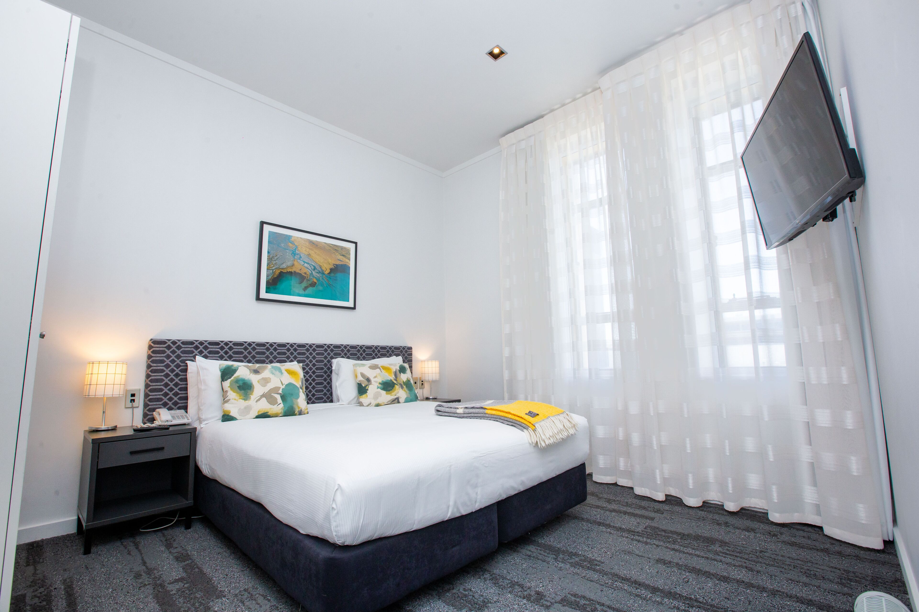 Een hotelkamer met een bed, nachtkastje, lamp, schilderij aan de muur en een aan de muur bevestigde televisie.