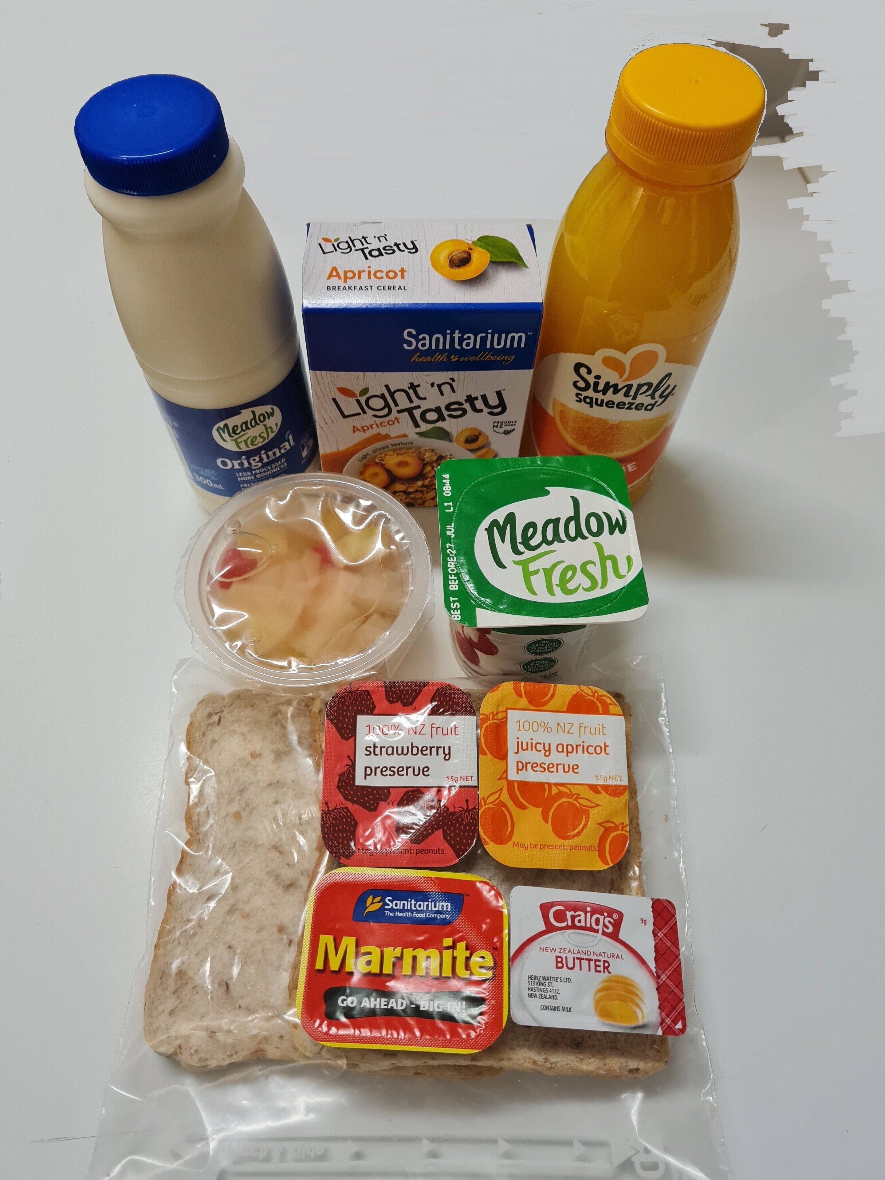 A variety of food items including a bottle of Meadow Fresh milk, a carton of Light 'n Tasty apricot cereal, a bottle of Simply Squeezed orange juice, a package of Marmite, and a packet of strawberry preserve.