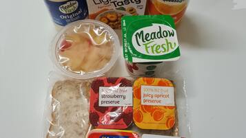 A variety of food items including a bottle of Meadow Fresh milk, a carton of Light 'n Tasty apricot cereal, a bottle of Simply Squeezed orange juice, a package of Marmite, and a packet of strawberry preserve.