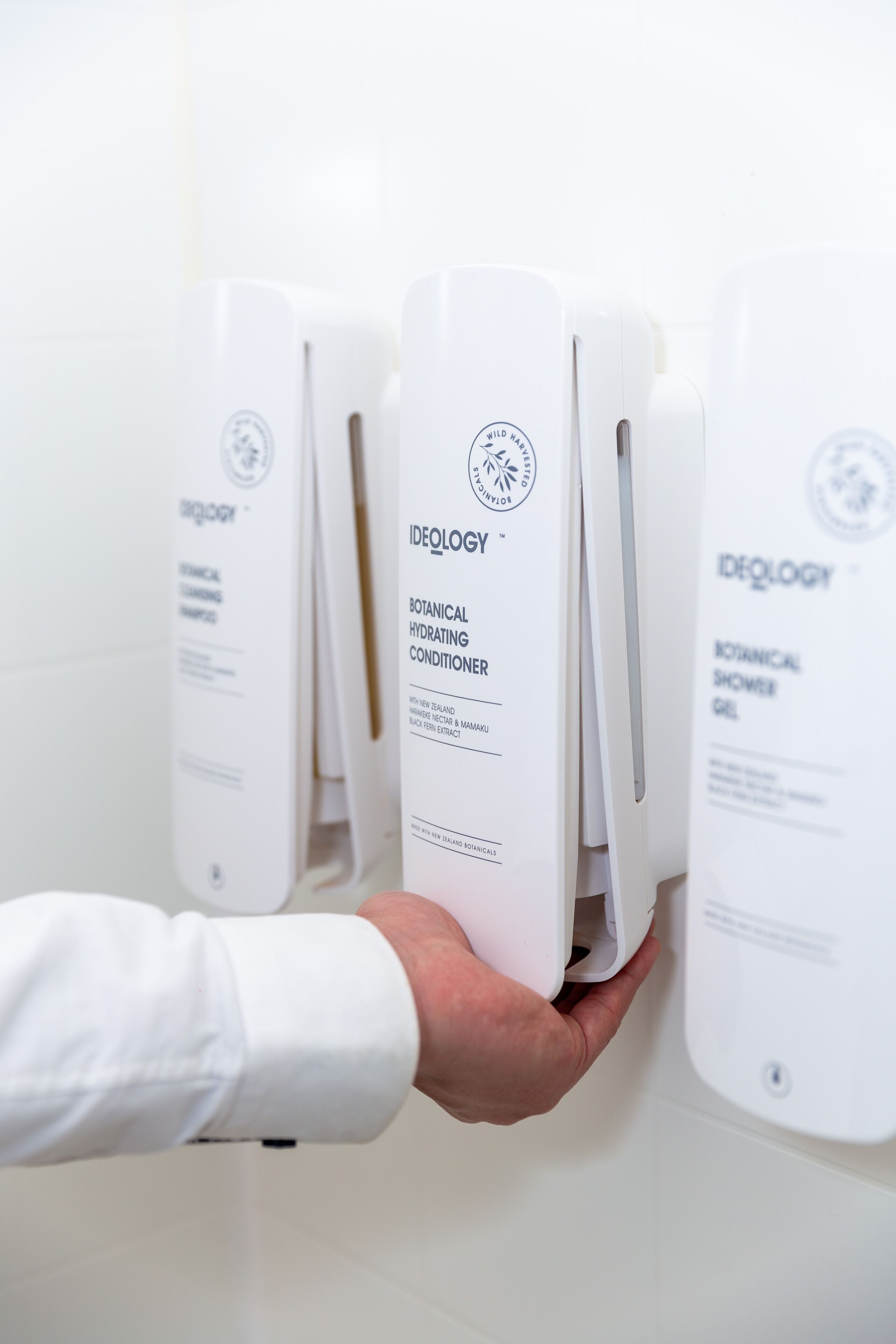 Una mano sostiene un dispensador blanco con la etiqueta 'Ideology Botanical Hydrating Conditioner'.