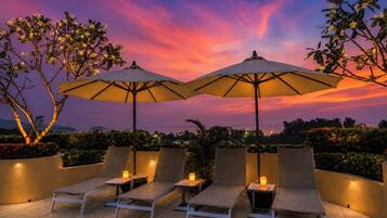 A terrace with lounge chairs and umbrellas, a sunset view, and a cityscape in the background.