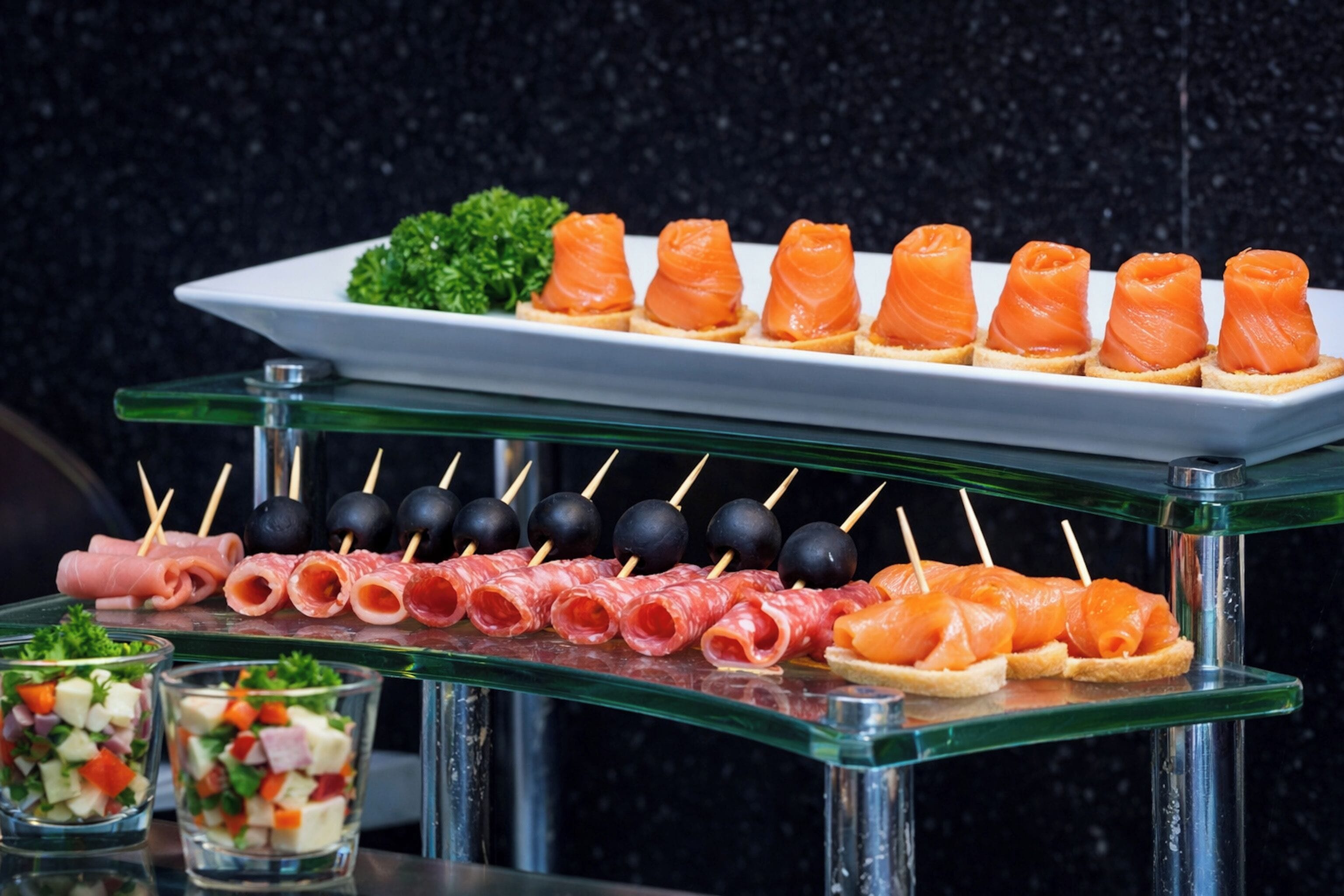 Un bufé con diversos alimentos, incluyendo salmón, prosciutto y una ensalada.