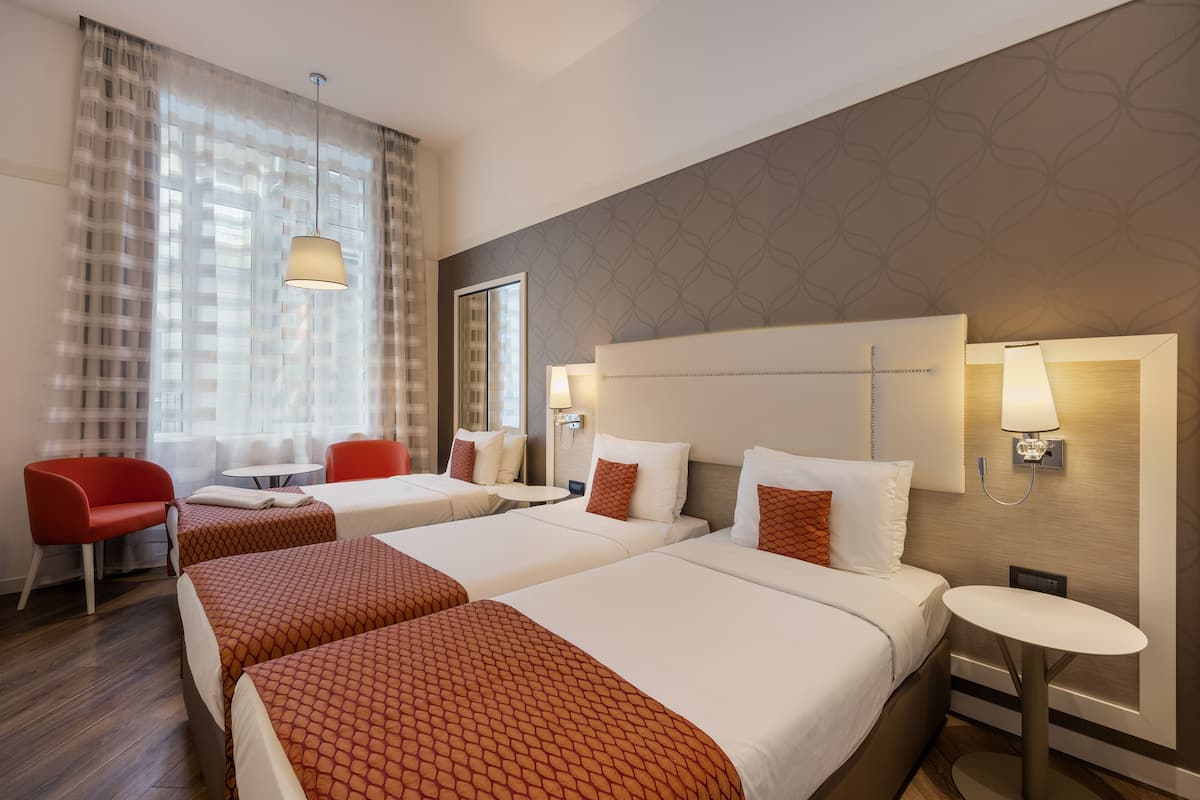 Una habitación de hotel con dos camas, una silla roja, una mesita pequeña y una lámpara de pared.