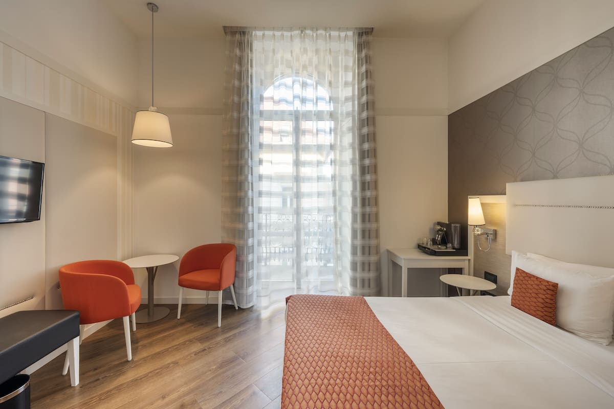 Habitación de hotel con una cama, dos sillones naranjas, una mesita, un televisor y una ventana con cortinas traslúcidas.