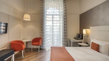 Una camera d'albergo con un letto, due poltrone arancioni, un tavolino, una televisione e una finestra con tende leggere.