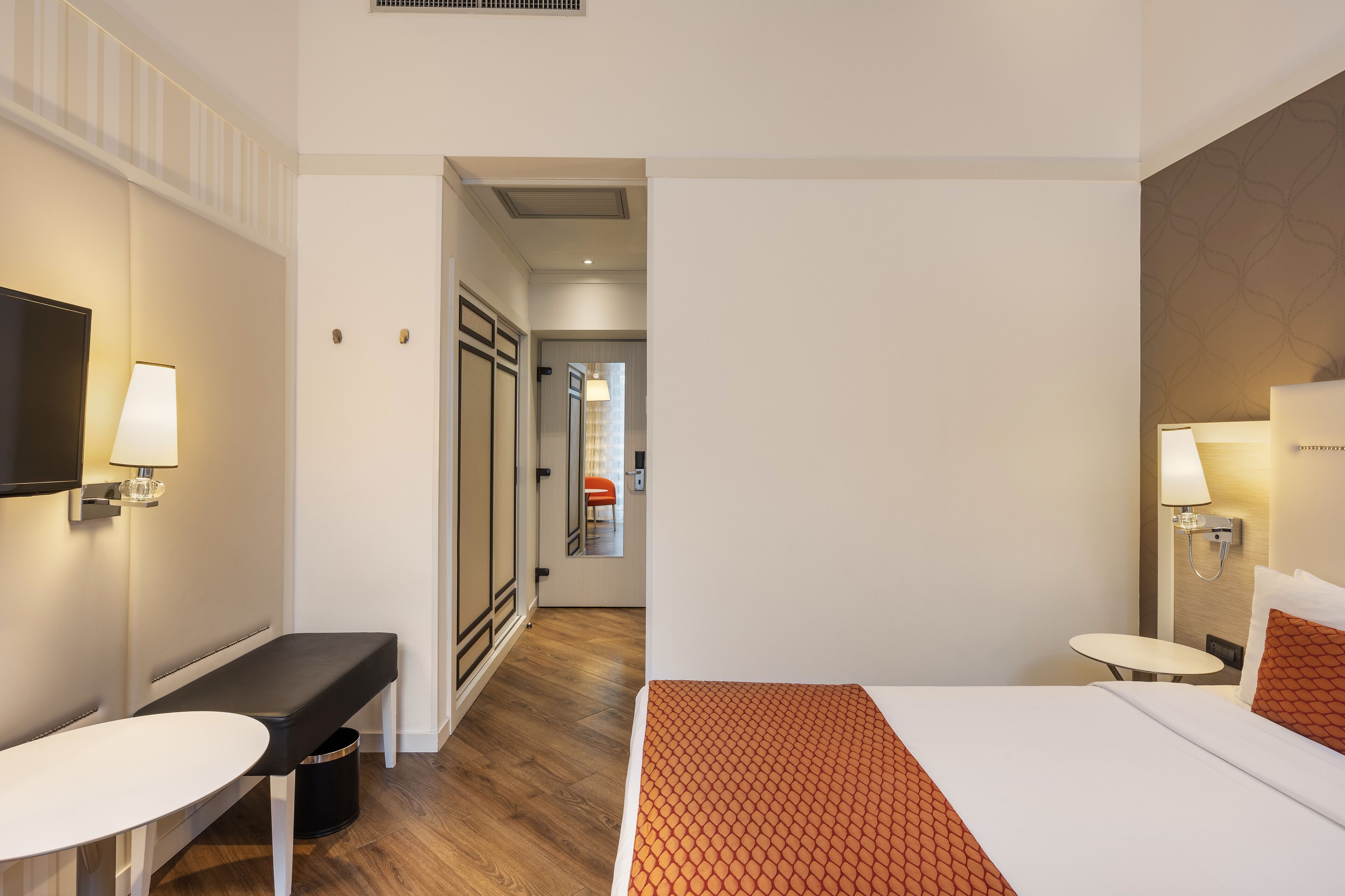 Una habitación de hotel con cama, mesita de noche, banca y televisor montado en la pared.