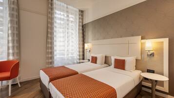 A hotel room with two beds, a small table, a chair, and a window with curtains.