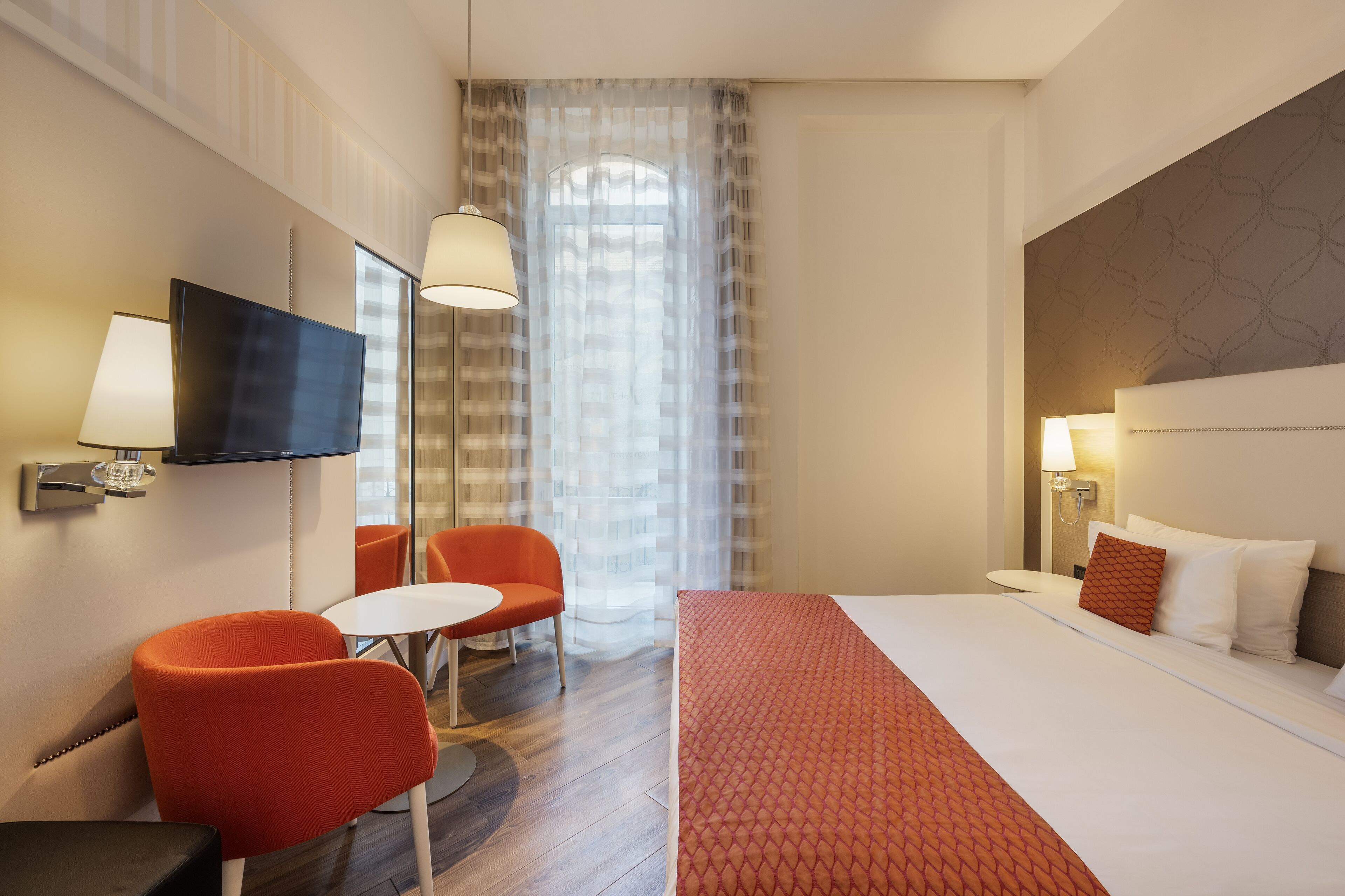 Habitación de hotel con cama, televisión, escritorio, dos sillas, ventana con cortinas y lámpara.