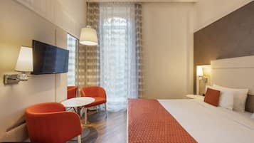 A hotel room with a bed, a TV, a desk, two chairs, a window with curtains, and a lamp.