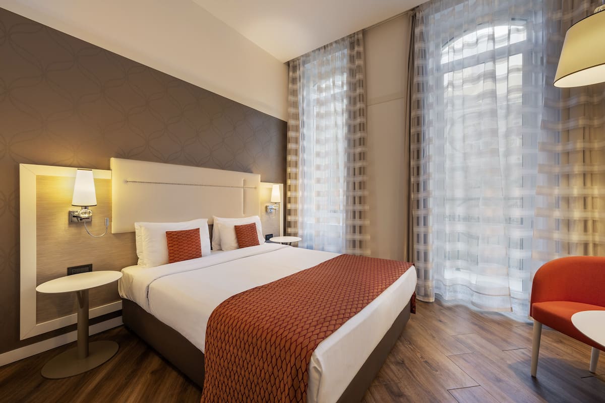 Una habitación de hotel con una cama grande, una mesita de noche, una silla y una lámpara.