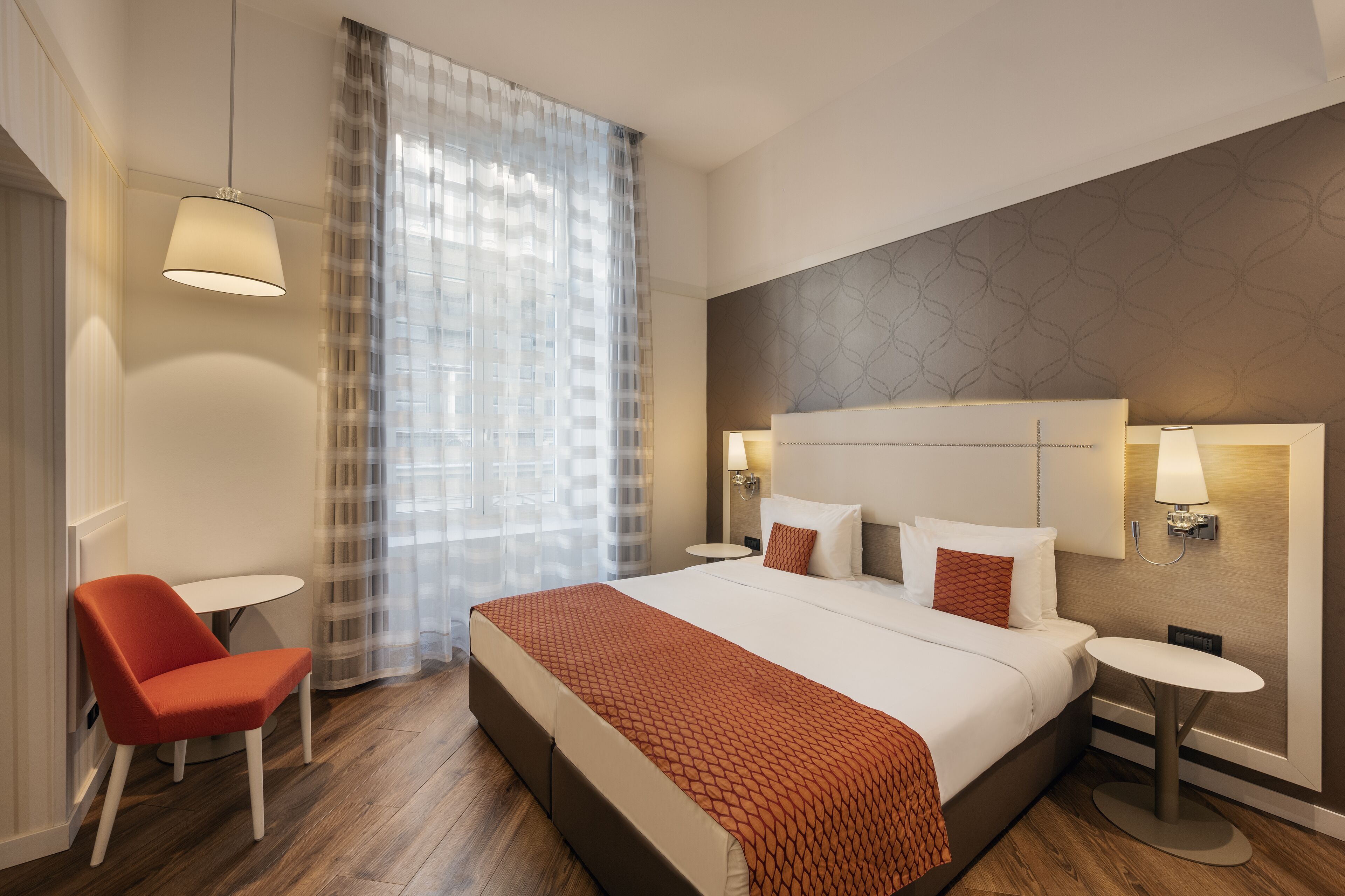 Une chambre d’hôtel moderne avec un grand lit, une chaise, une petite table et une fenêtre avec des rideaux légers.