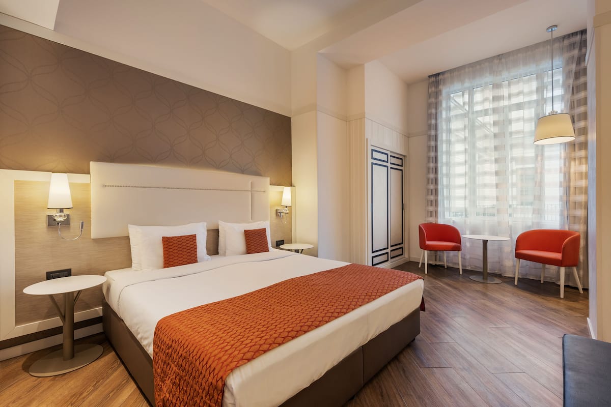 Una habitación de hotel con una cama grande, dos mesitas de noche, una mesa redonda pequeña, un sofá rojo y un ventanal con cortinas.