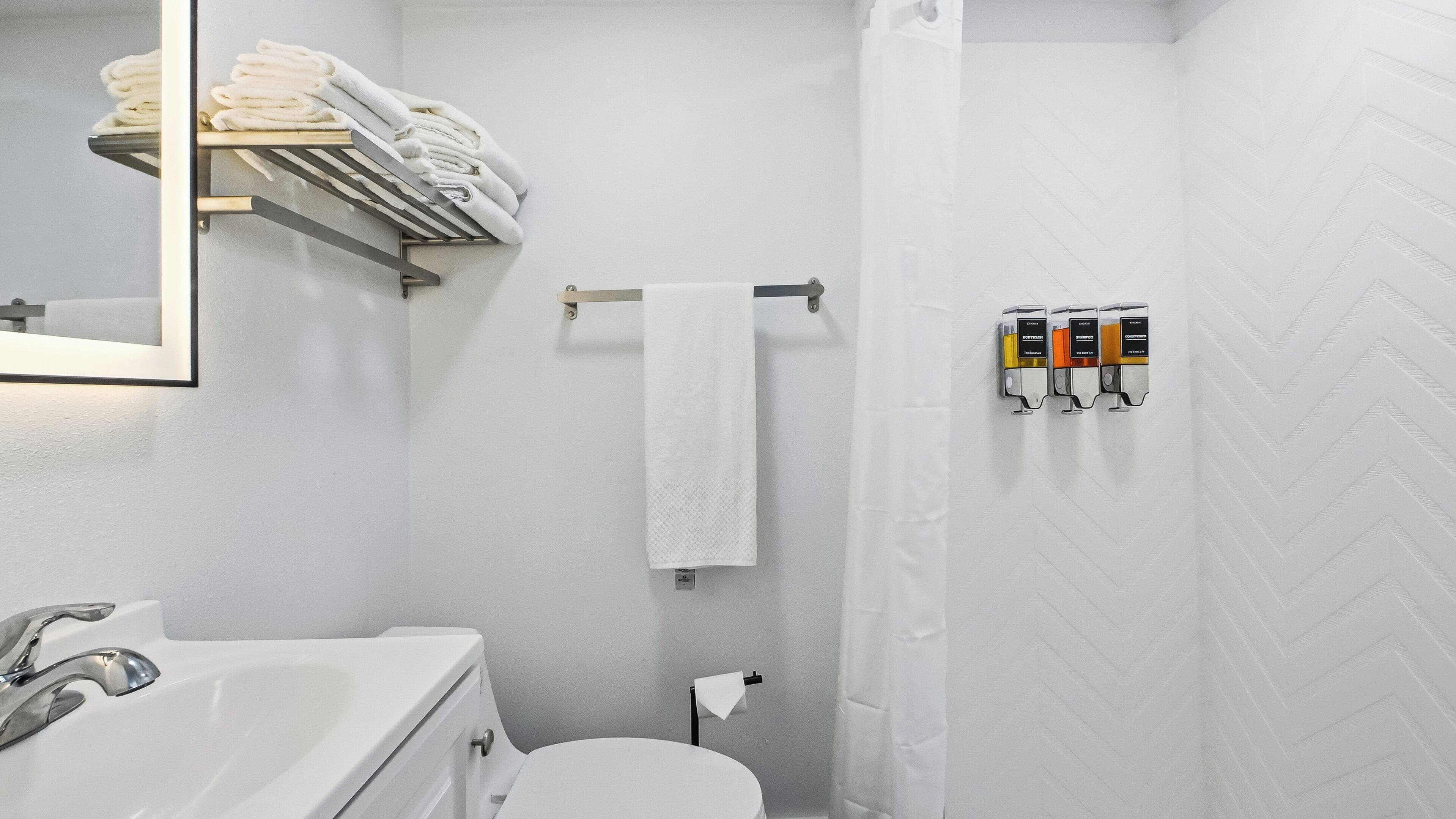 A bathroom with a white sink, a mirror, a towel rack with folded towels, a toilet, and a shower curtain.