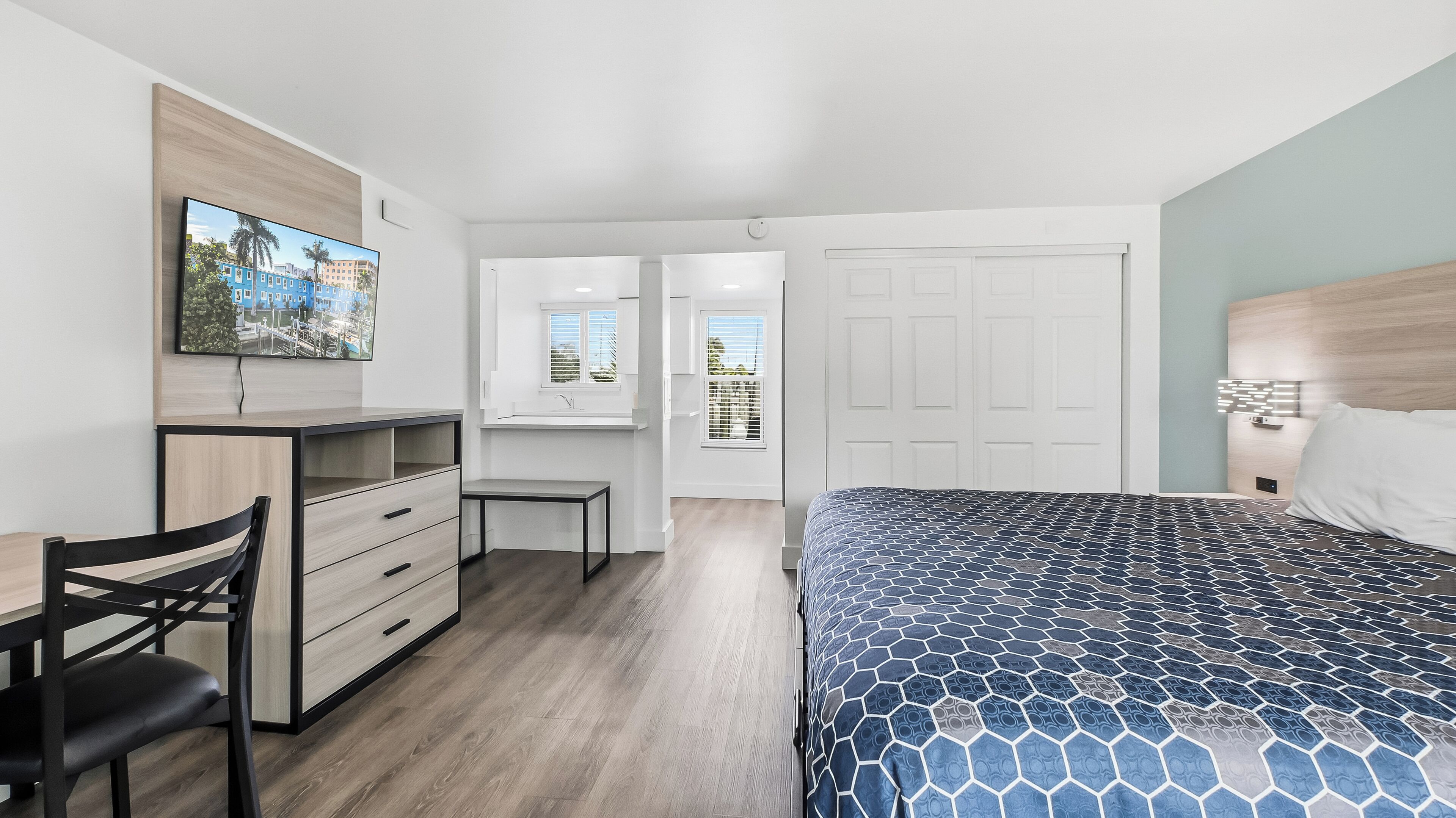 Een moderne slaapkamer met een bed, een bureau en een stoel. Er is een raam met zonwering, een deur en een aan de muur bevestigde televisie.