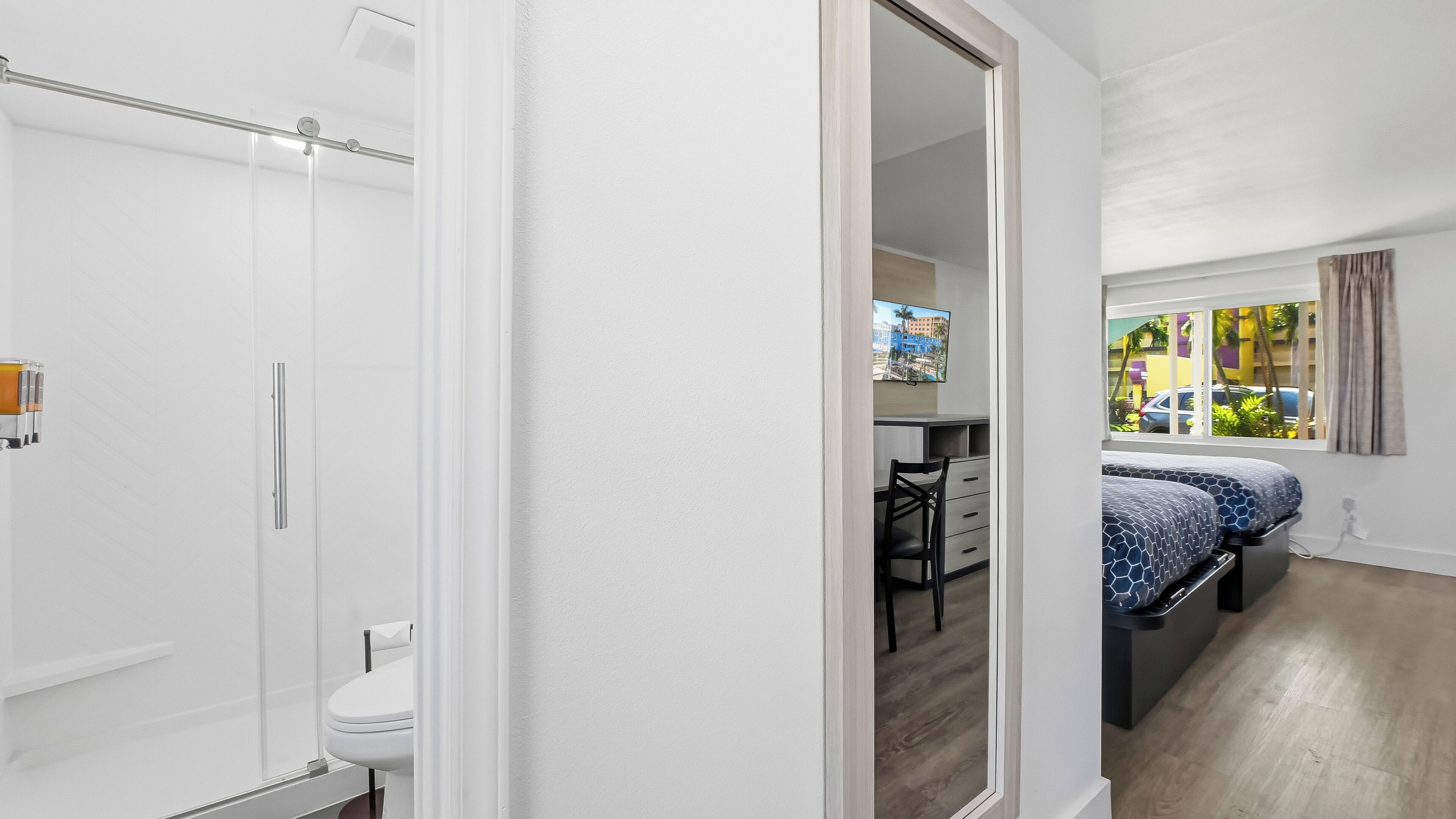 A modern bathroom with a shower, toilet, and a glass door leading to a bedroom with a bed, desk, and window with a view of buildings and greenery.