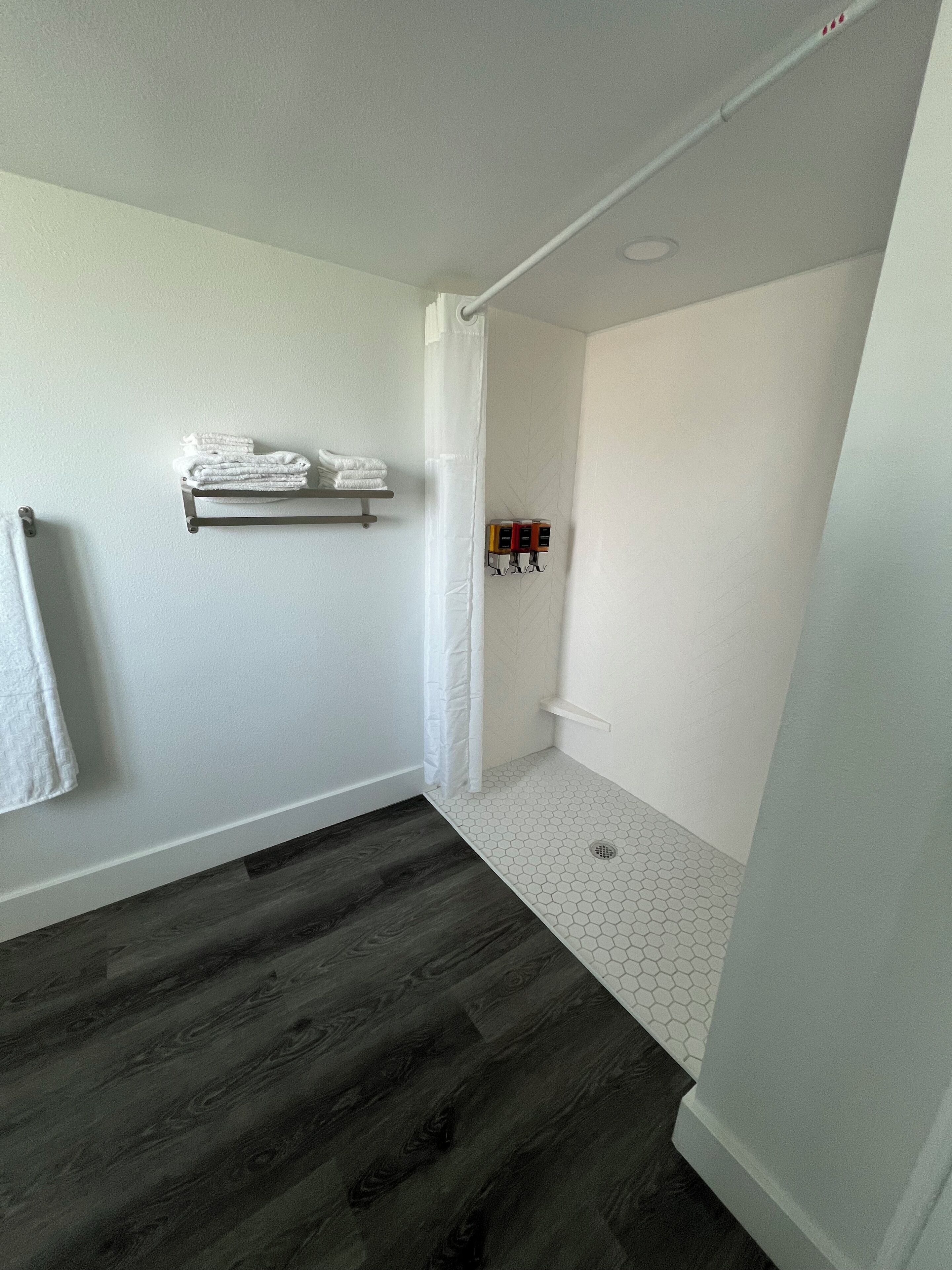 Une salle de bain avec un plancher en bois, un porte-serviettes avec des serviettes pliées, un rideau de douche et une étagère contenant des articles de toilette.
