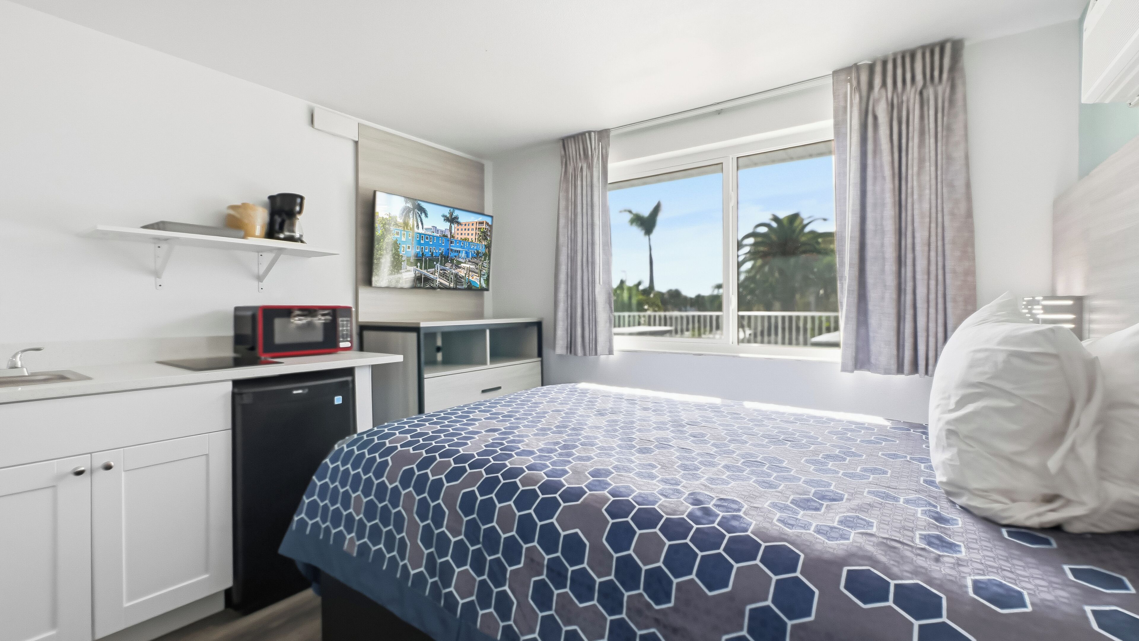 Une chambre d’hôtel avec un lit, une petite kitchenette, une fenêtre donnant sur des palmiers et un tableau au mur.