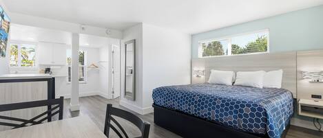 A modern hotel room with a bed, a desk, and a chair. There is a window with a view of palm trees.