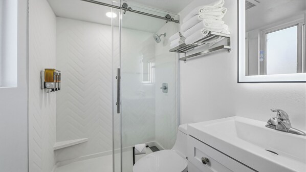 Une salle de bain moderne avec une douche à parois de verre, un lavabo blanc avec un miroir et des serviettes soigneusement pliées et rangées sur une étagère.