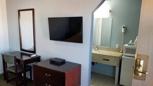 Una habitación de hotel con escritorio, silla, cómoda, televisor y baño que incluye lavamanos, espejo y retrete.
