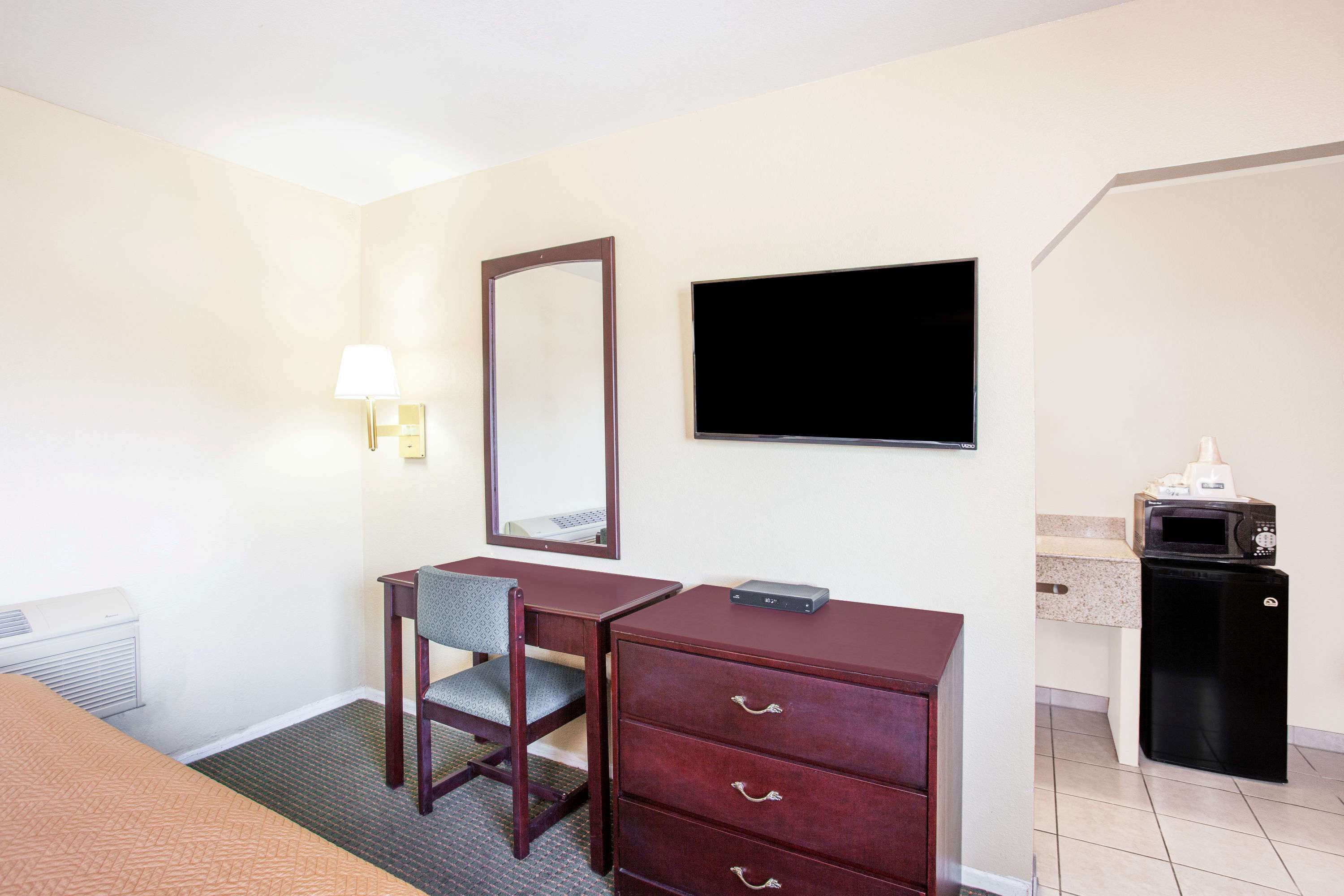 A hotel room with a bed, a desk with drawers, a chair, a flat-screen TV, a mirror, a lamp, and a small kitchenette with a microwave.