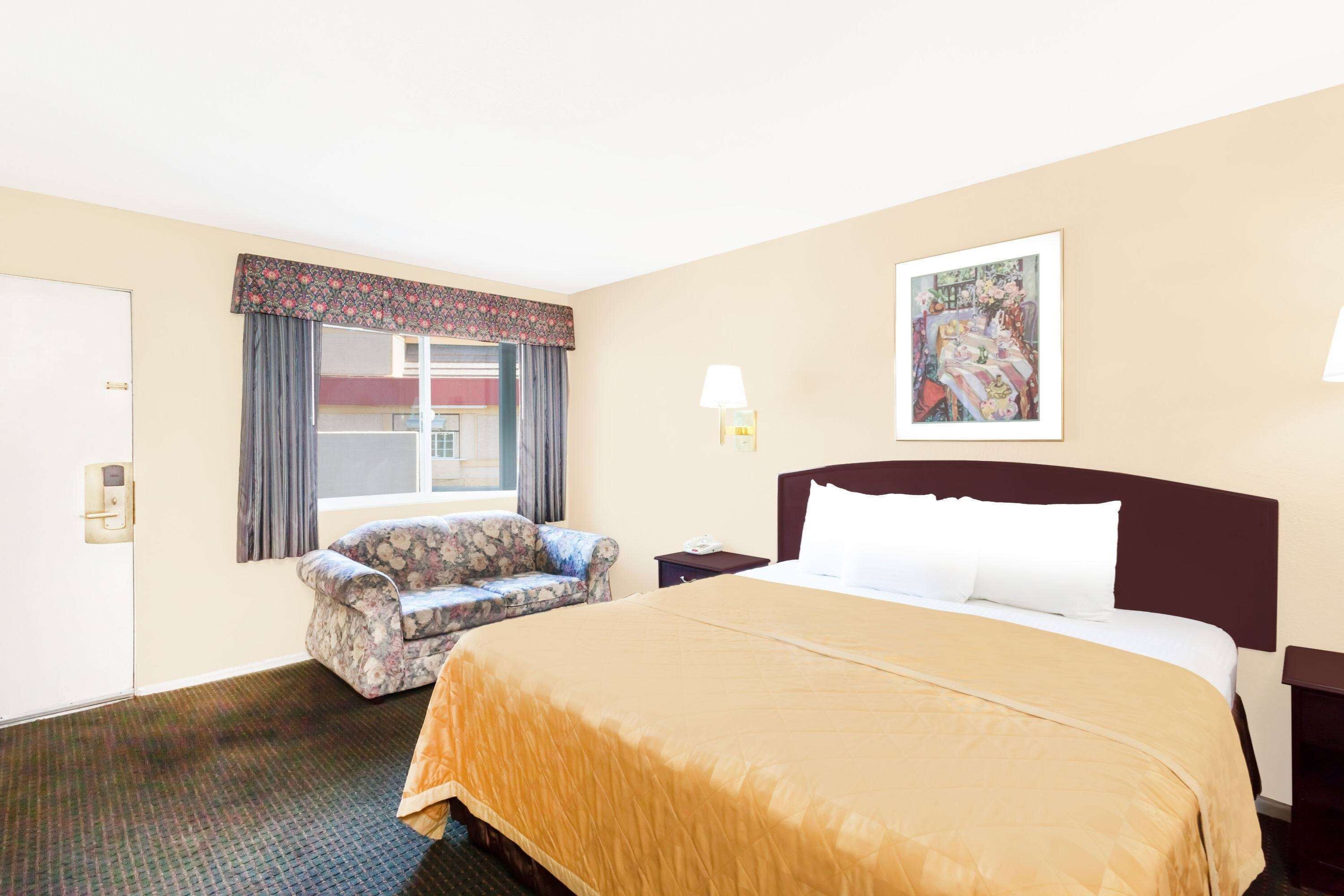 A hotel room with a large bed, a chair, a desk, a lamp, a painting on the wall, and a window with curtains.