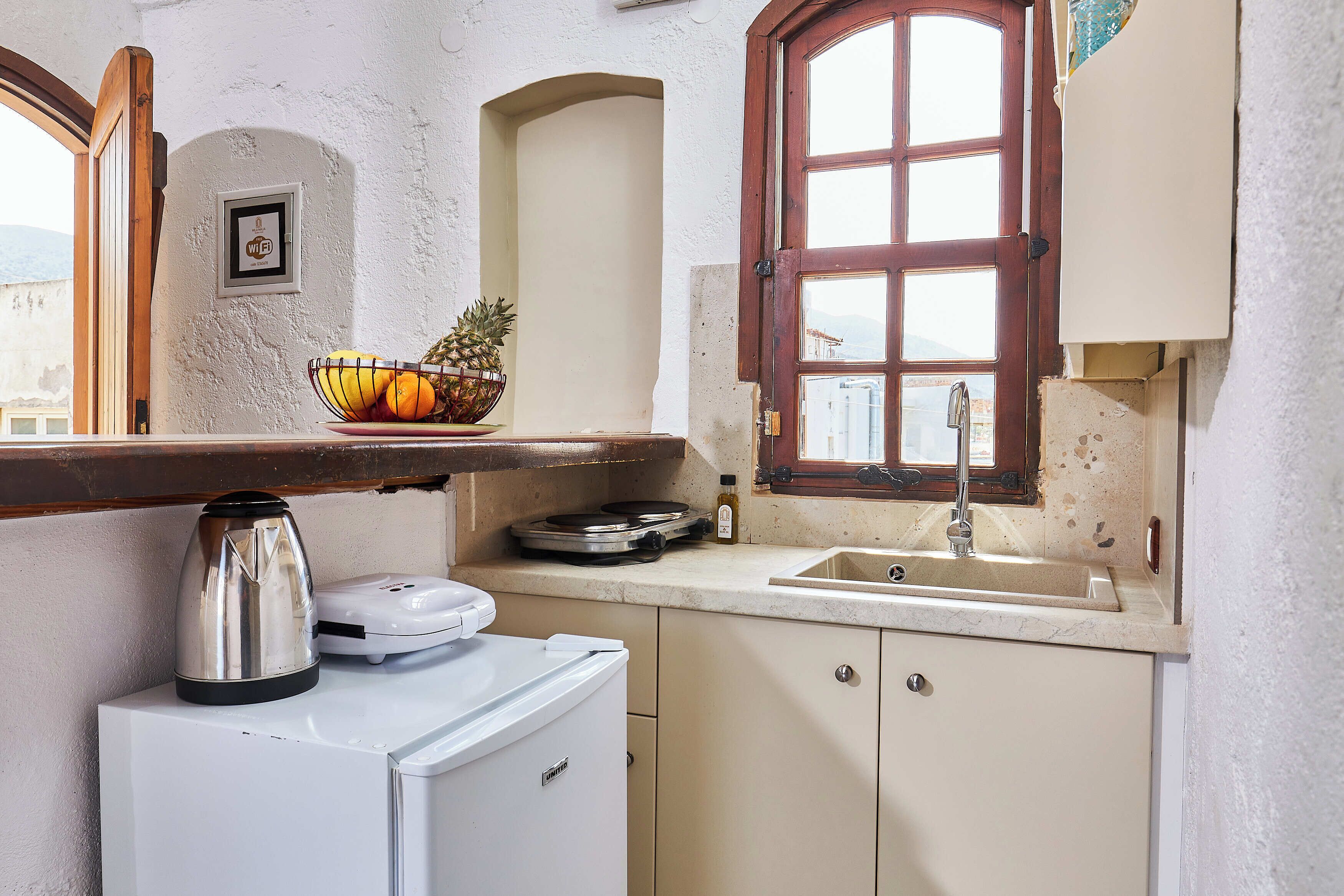 Cozinha compacta com geladeira branca, cafeteira, fruteira e janela com vista.
