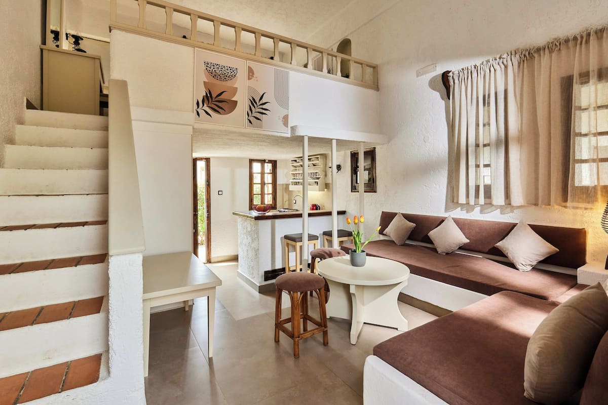 Een ruime woonkamer met een hoge plafond, een trap, een eettafel met krukken en een zithoek.