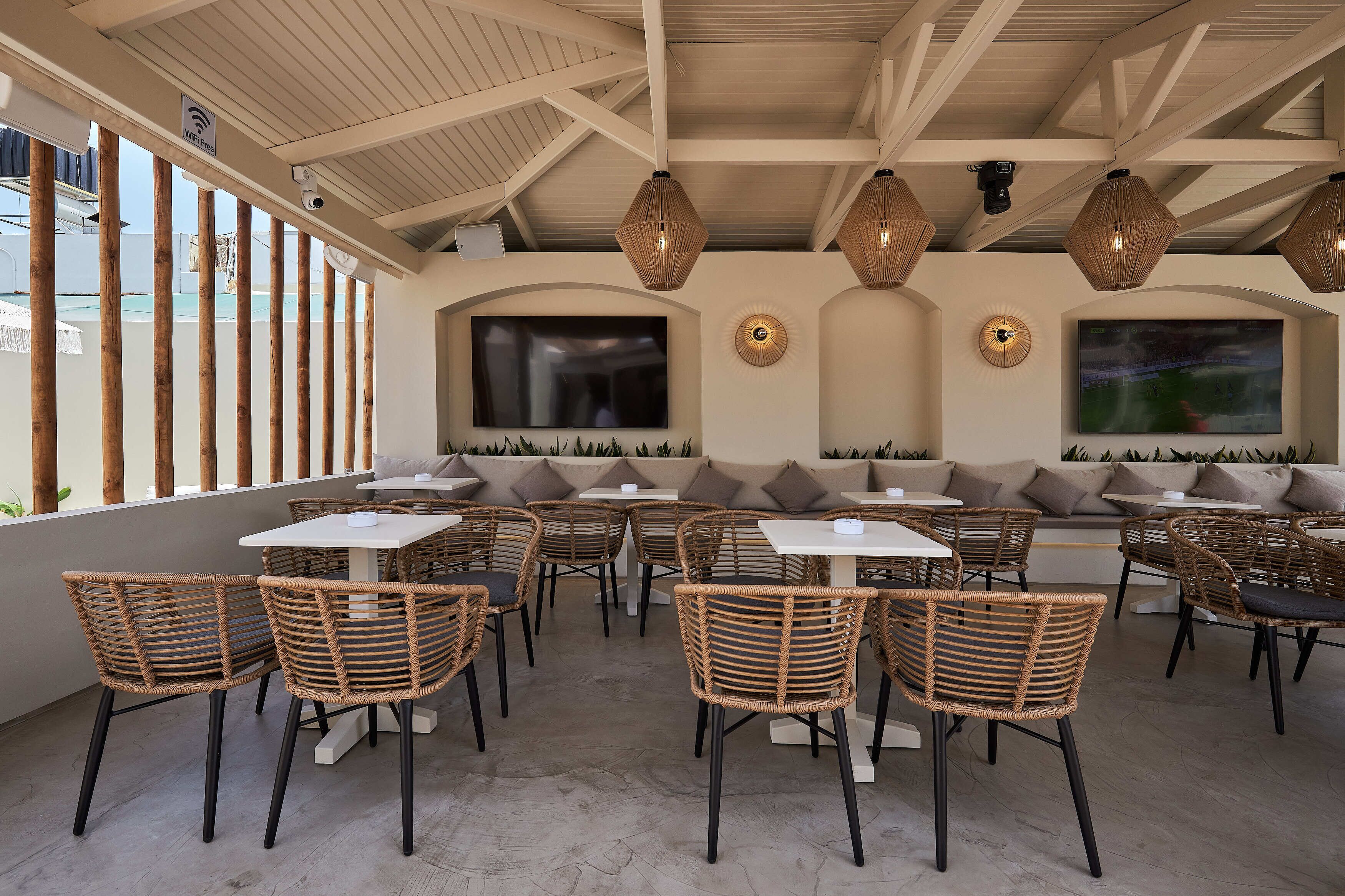 A café with wicker chairs and tables, a TV mounted on the wall, and a ceiling with exposed beams.