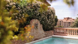 Una piscina con pared de ladrillo y barandilla, rodeada de abundante vegetación y un edificio al fondo.