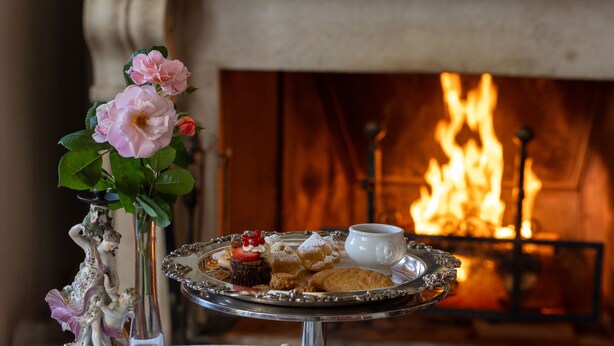 Una mesa dispuesta para el té, con una bandeja de pasteles, un florero y una chimenea al fondo.