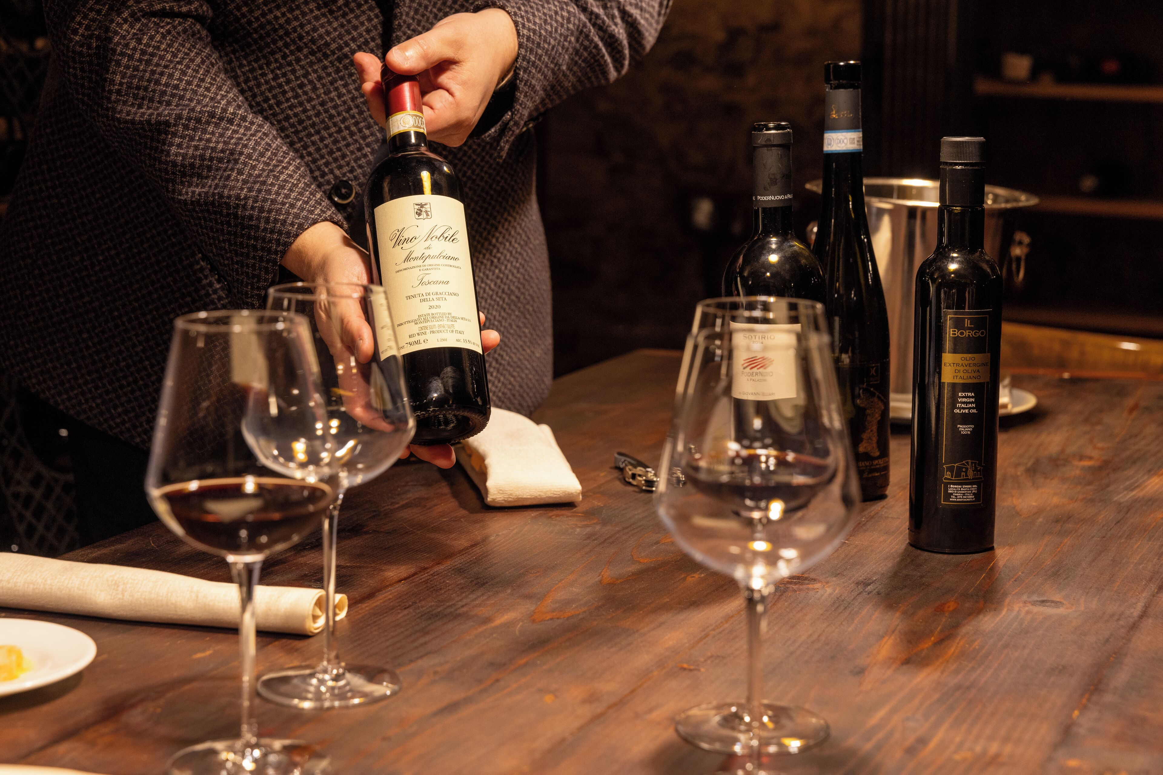 A person pouring wine into a glass on a wooden table with several wine bottles and glasses.