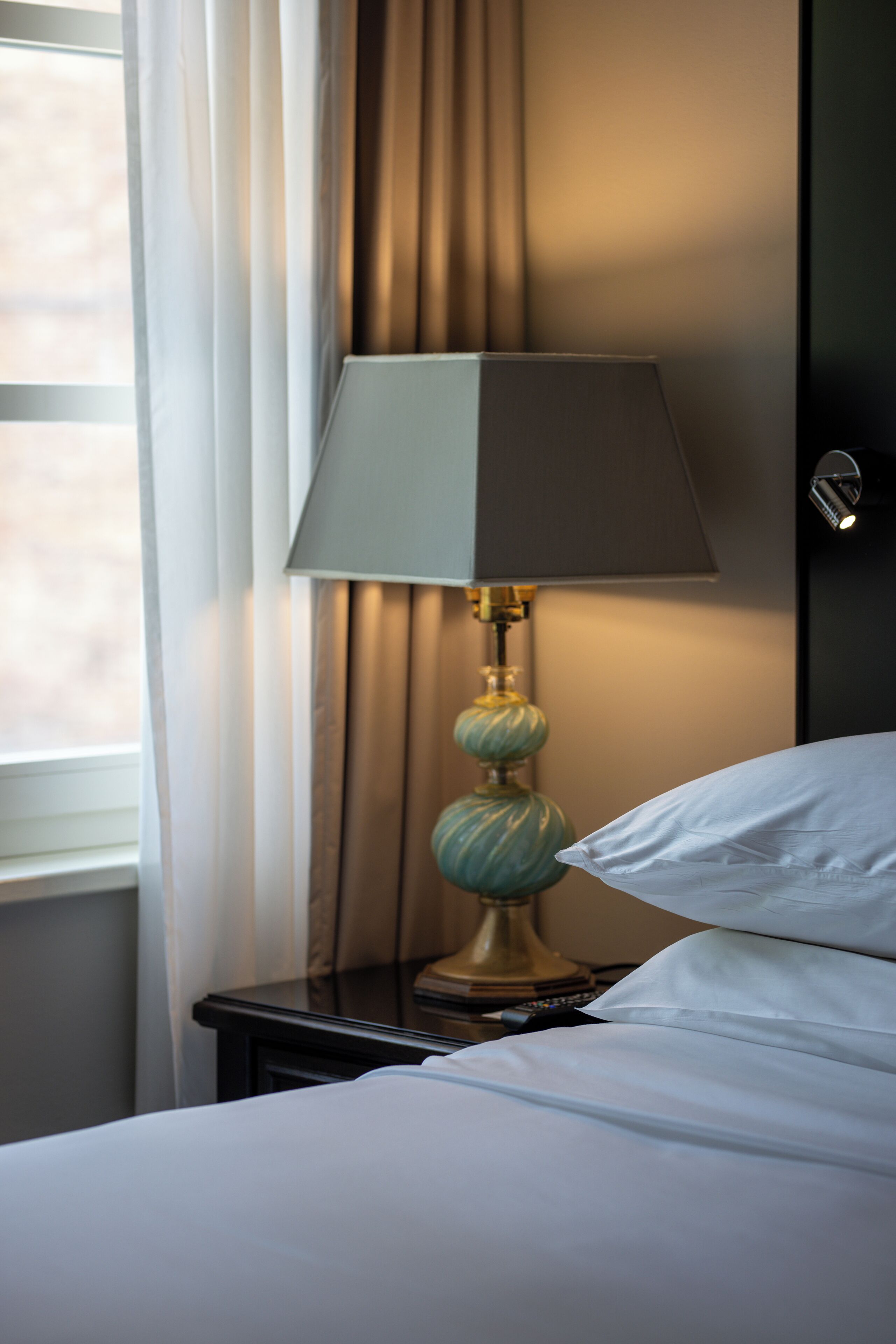 Ein Hotelzimmer mit Bett, Nachttisch, Lampe und einem Fenster mit Vorhängen.