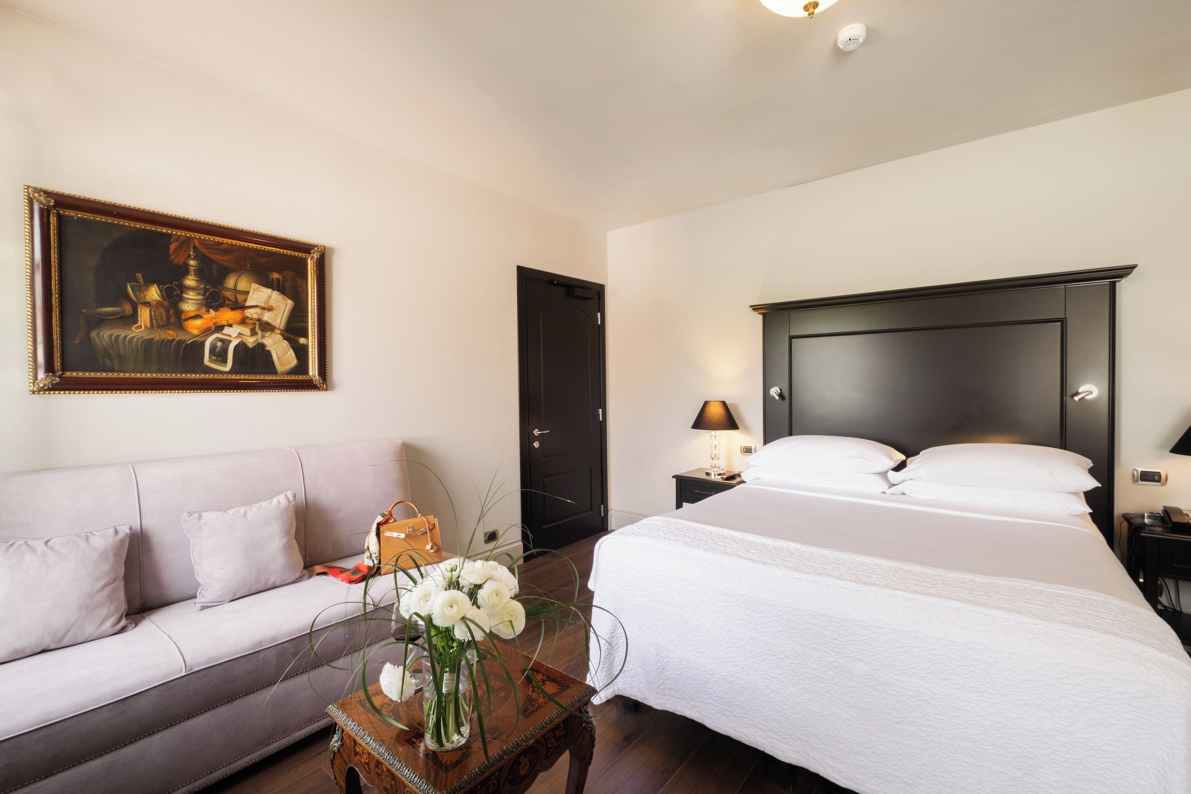 Habitación de hotel con una cama grande, un sofá, un cuadro en la pared y un florero con flores sobre una mesita de noche.
