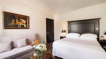 Habitación de hotel con una cama grande, un sofá, un cuadro en la pared y un florero con flores sobre una mesita de noche.