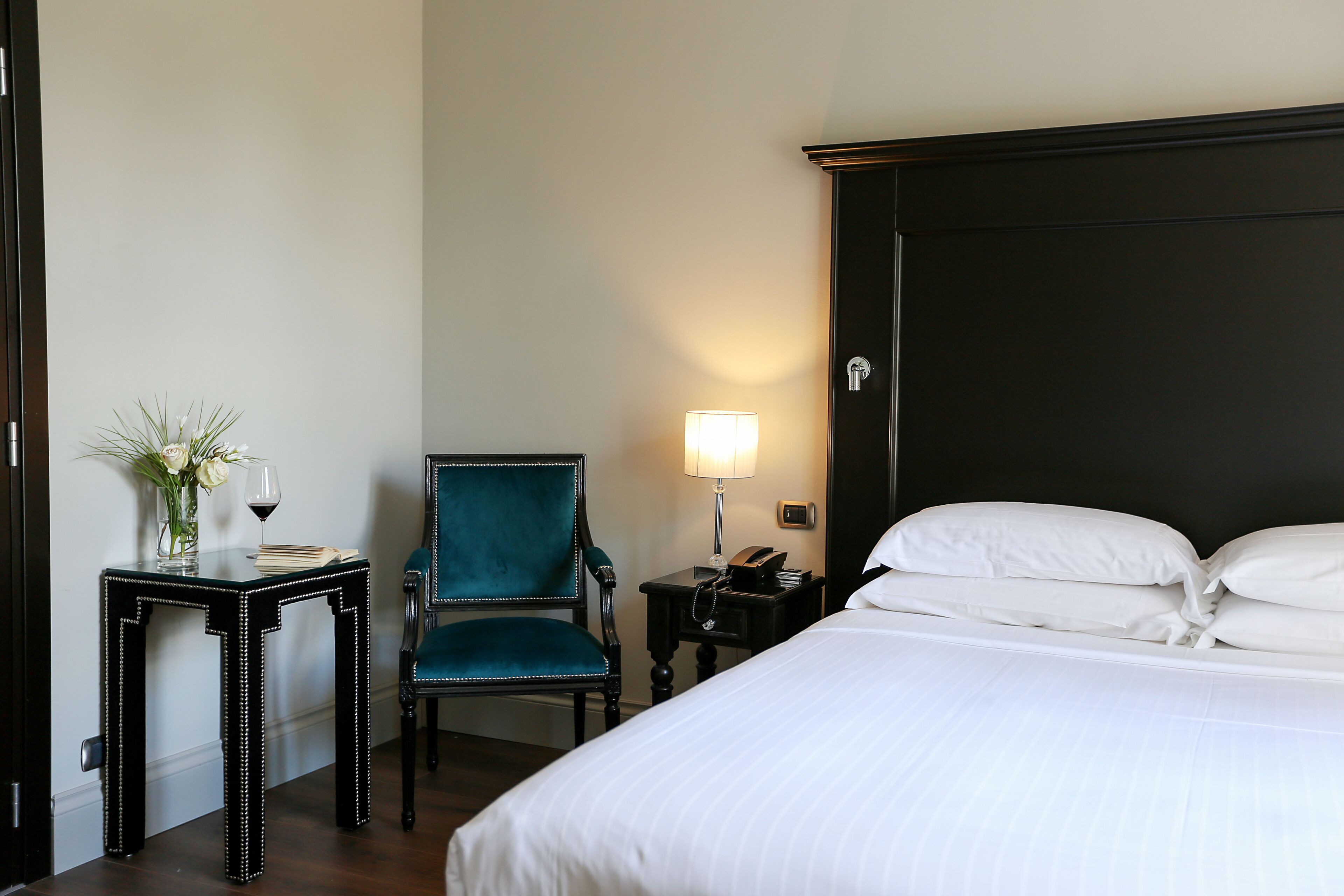A hotel room with a bed, a chair, a table with a vase and wine, and a nightstand with a lamp.