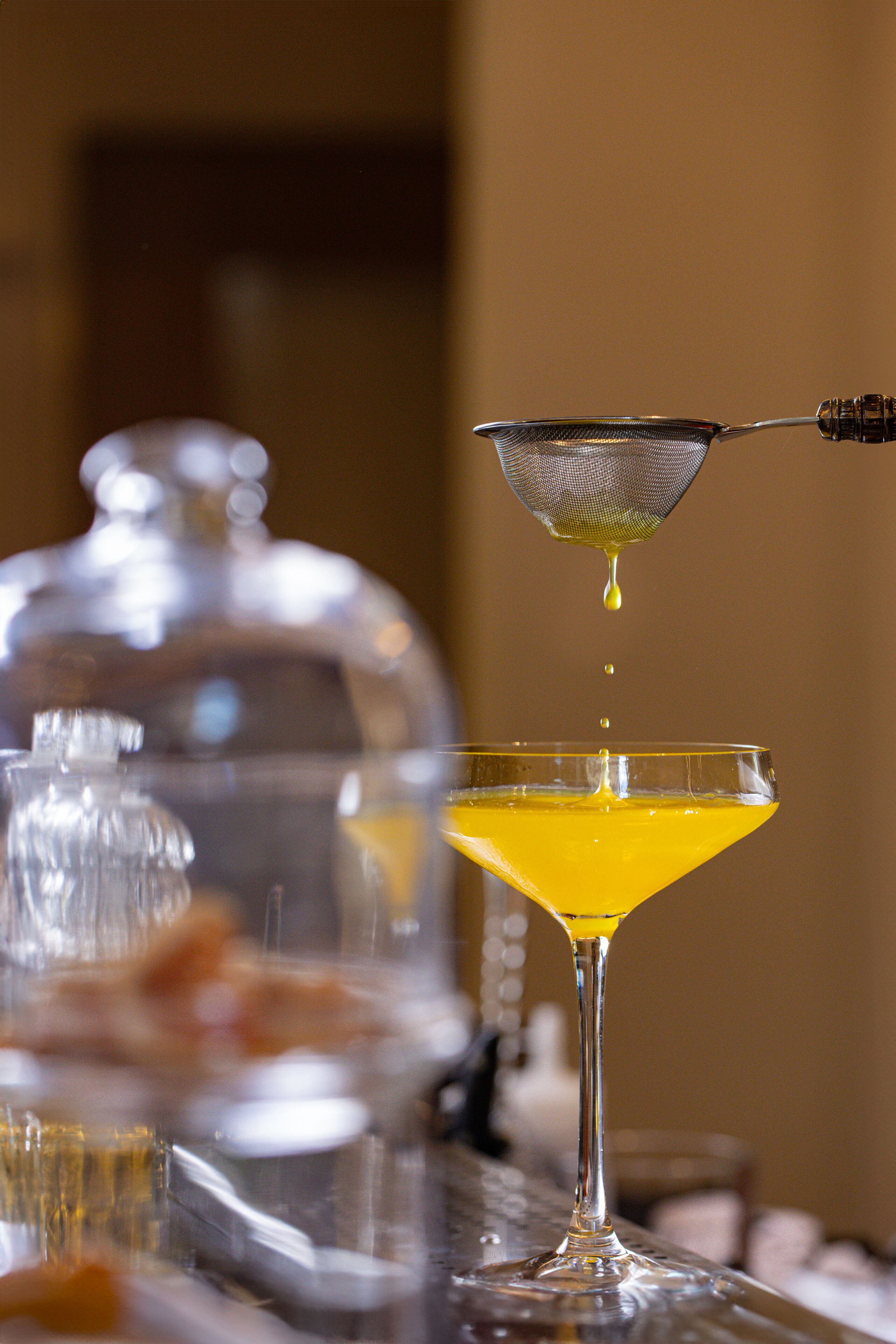 Una copa de martini con un líquido amarillo, un colador y un recipiente de vidrio al fondo.