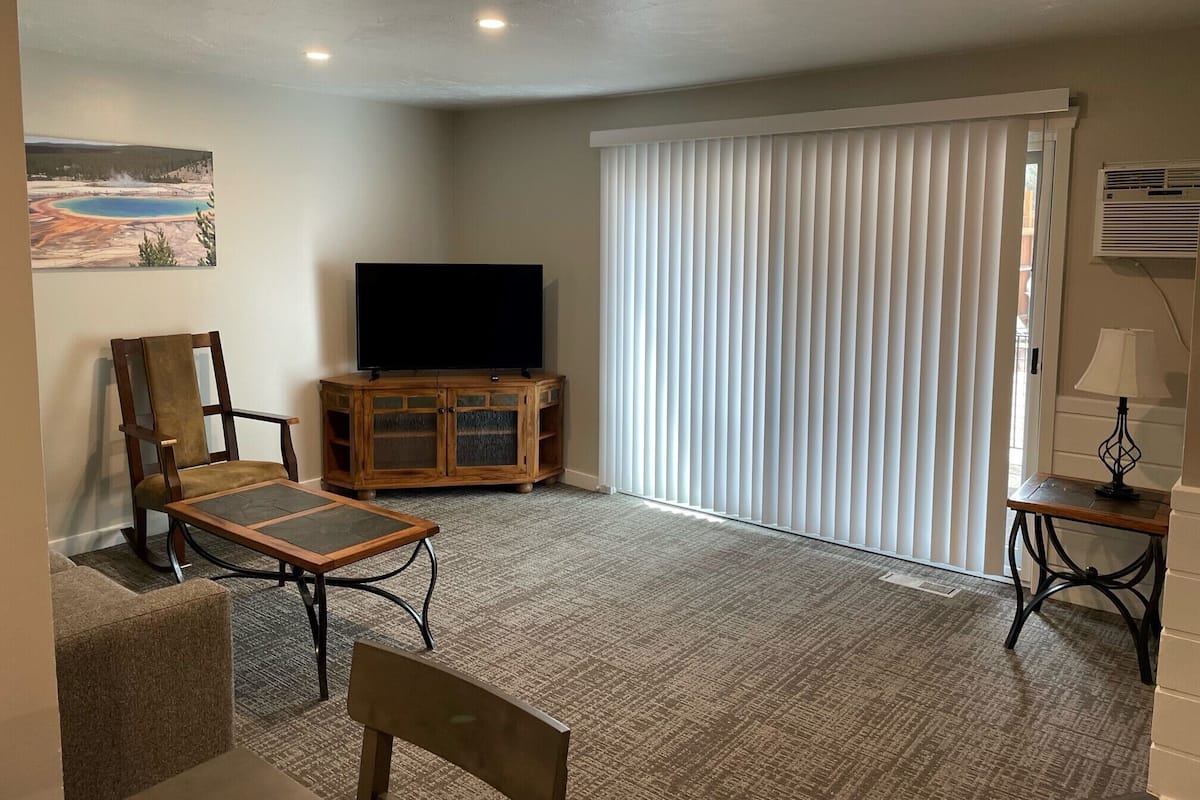A living room with a flat-screen TV on a wooden cabinet, a cushioned chair, a small table with a lamp, and a large window with blinds.