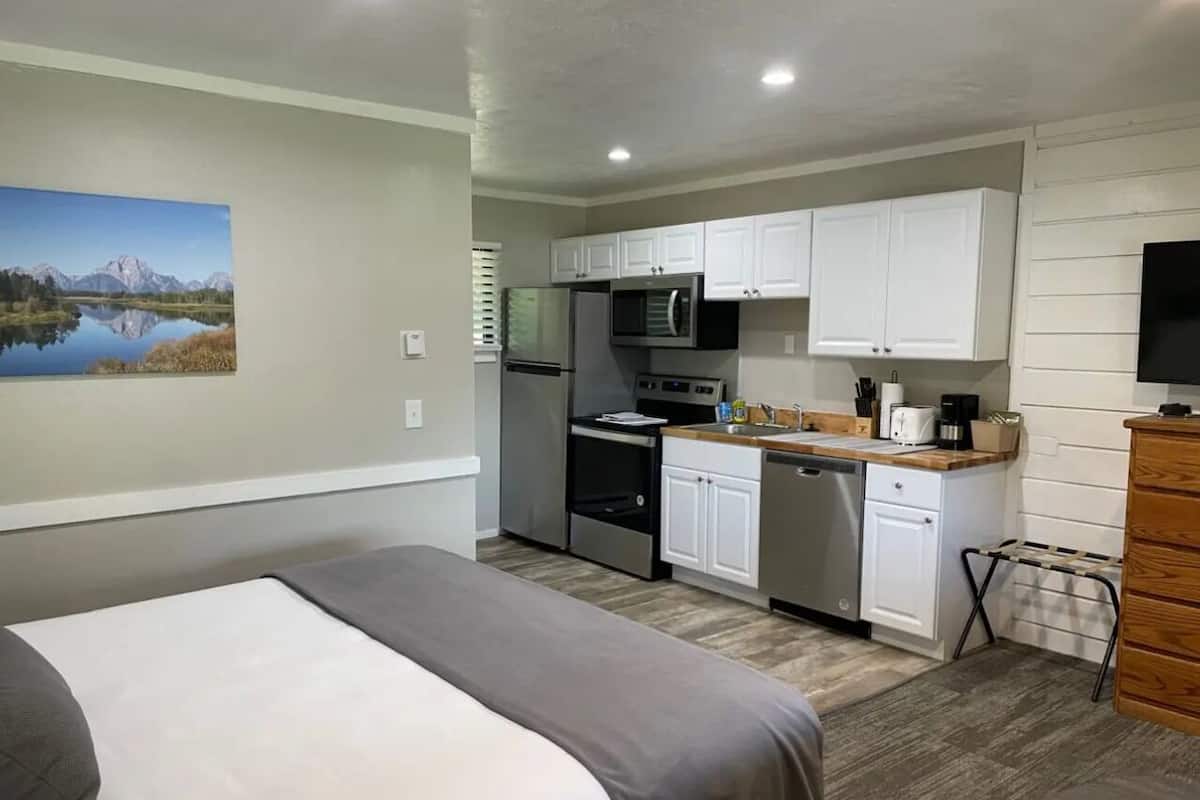 A compact hotel room with a bed, kitchenette, and a painting of a mountain lake.