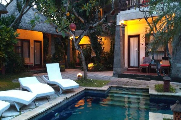 Uma área de piscina com espreguiçadeiras, uma piscina pequena e um edifício com telhado de palha.