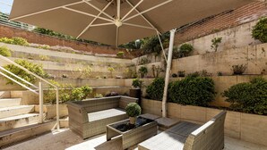 A sunlit outdoor seating area with wicker furniture, a large umbrella, and a brick wall with plants. - Wall Art Hotel (Prato)