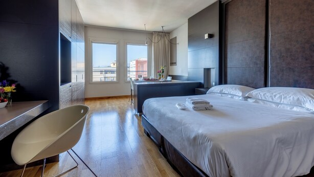 A modern hotel room with a large bed, a desk, a chair, and a view of the city through large windows. - Wall Art Hotel (Prato)