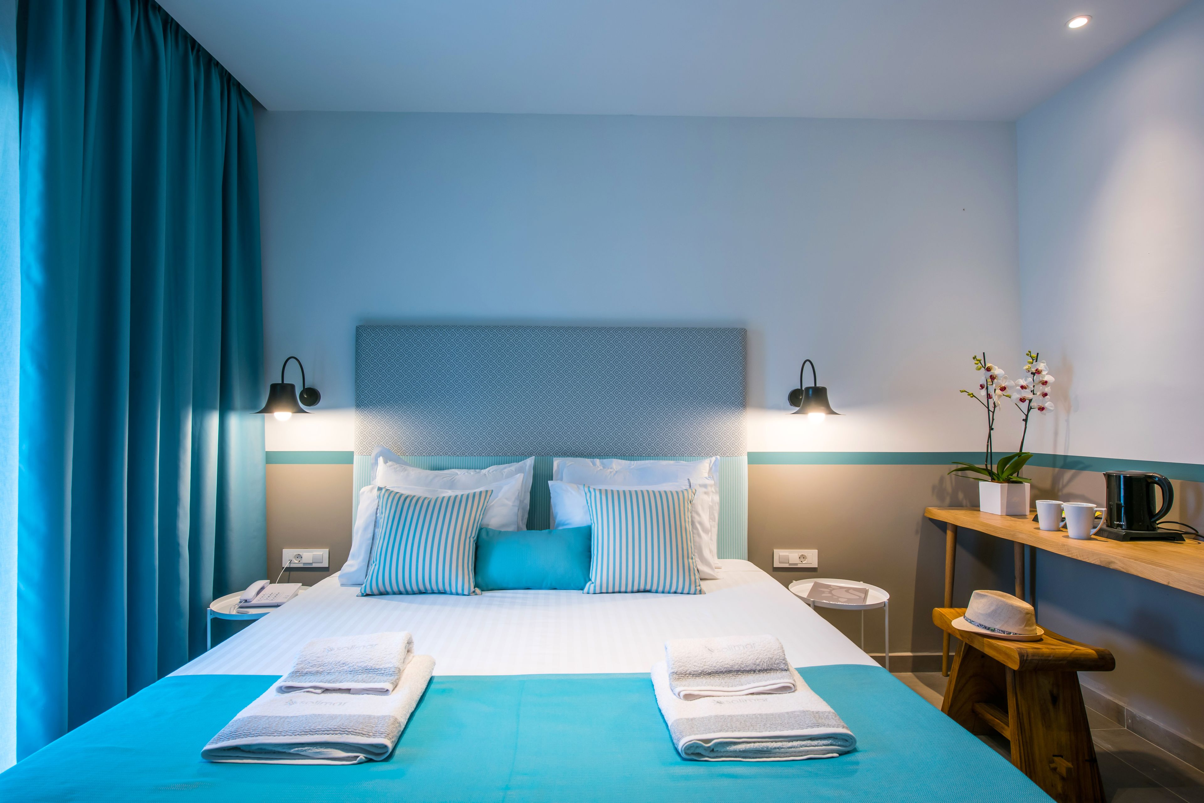 Une chambre d’hôtel moderne avec un grand lit, deux tables de chevet, un petit tabouret, une commode et une étagère murale agrémentée d’une plante et d’une machine à café.