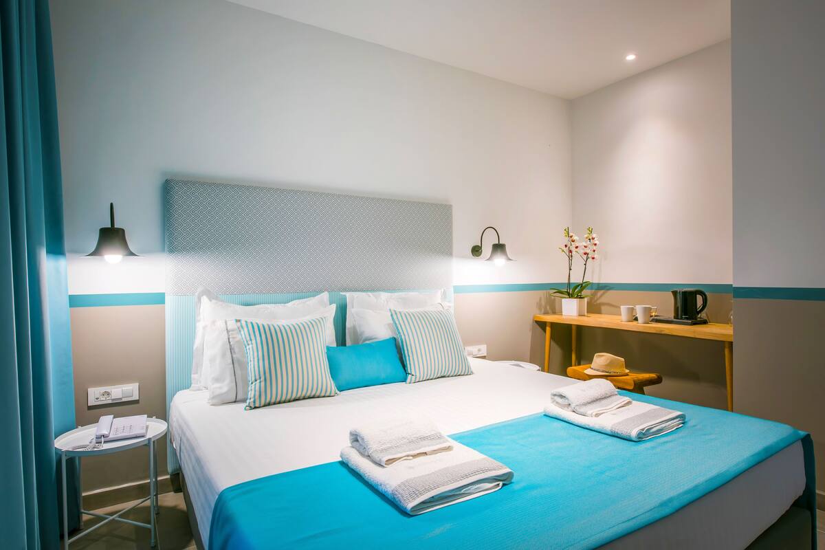 A neatly made bed with blue bedding, a bedside table with a phone, and a nightstand with a lamp.