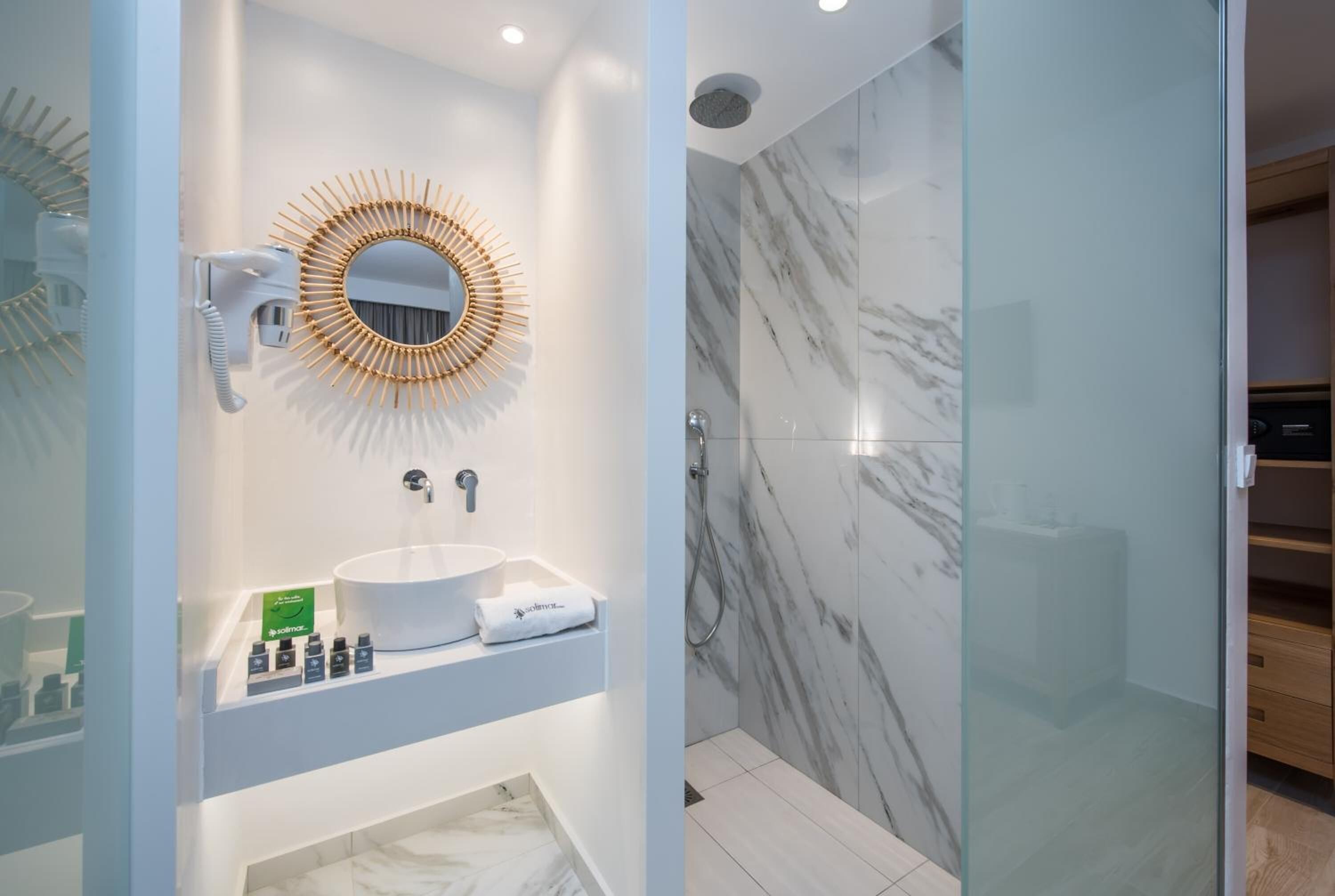 A modern bathroom with a white sink, a round mirror with a sunburst frame, a handheld hair dryer, and a glass-enclosed shower area with marble walls.