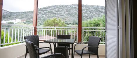 A balcony with a table and chairs, a view of a hilly landscape, and a white railing.