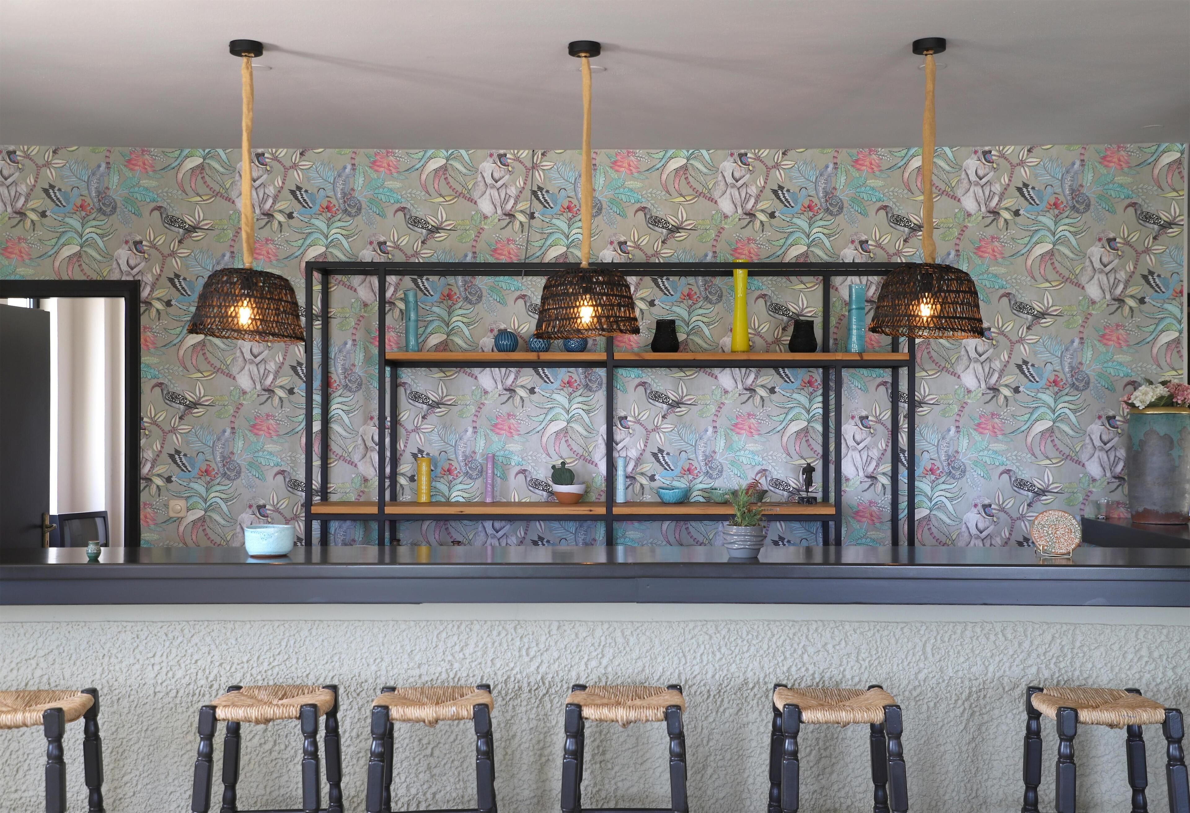 Un coin bar agrémenté d’un papier peint à motifs, d’une étagère en bois ornée d’objets décoratifs et de tabourets de bar en osier.